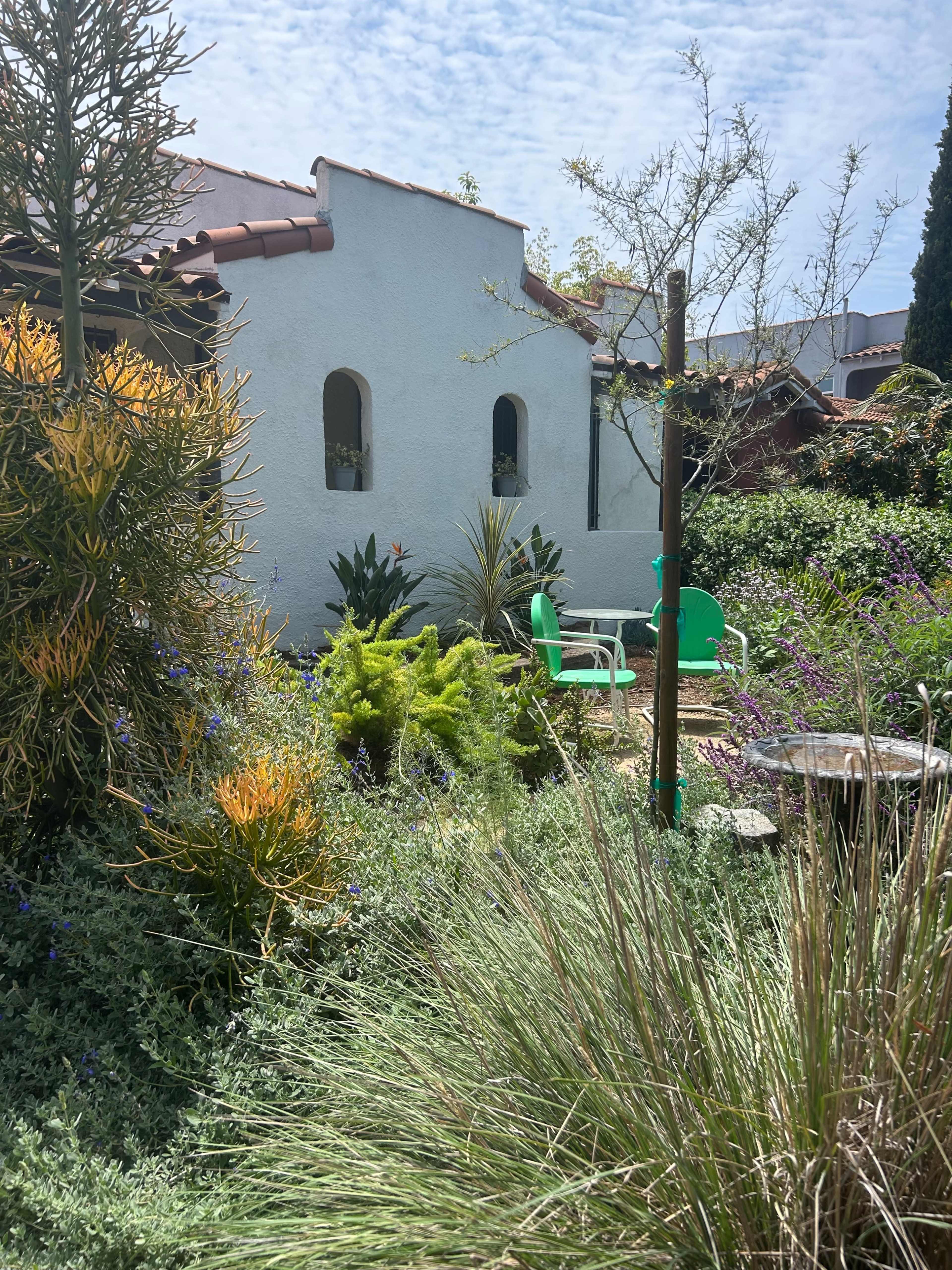 The image shows a tranquil garden with vibrant greenery, a small path leading to two green chairs, and a white stucco building in the background.