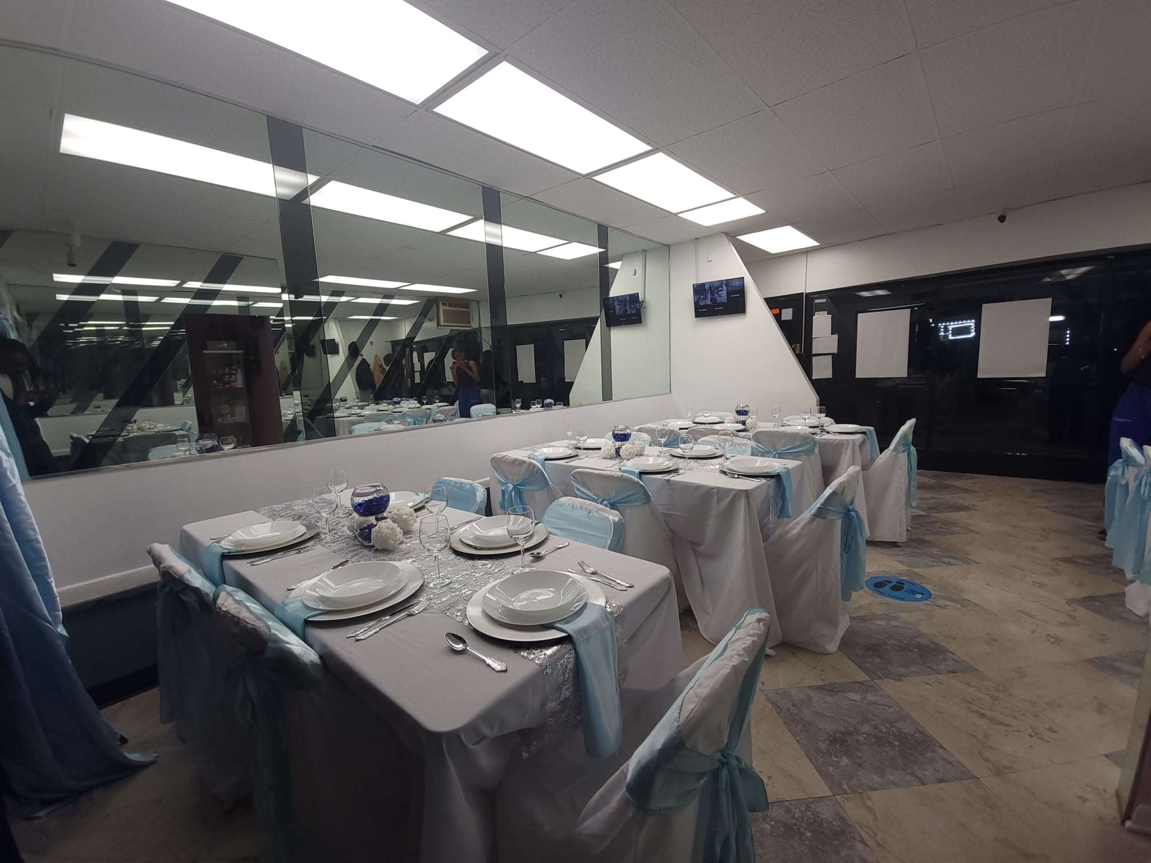 A dining area is set with multiple tables covered in white tablecloths and blue accents, arranged for a formal meal, with mirrored walls reflecting the setup.