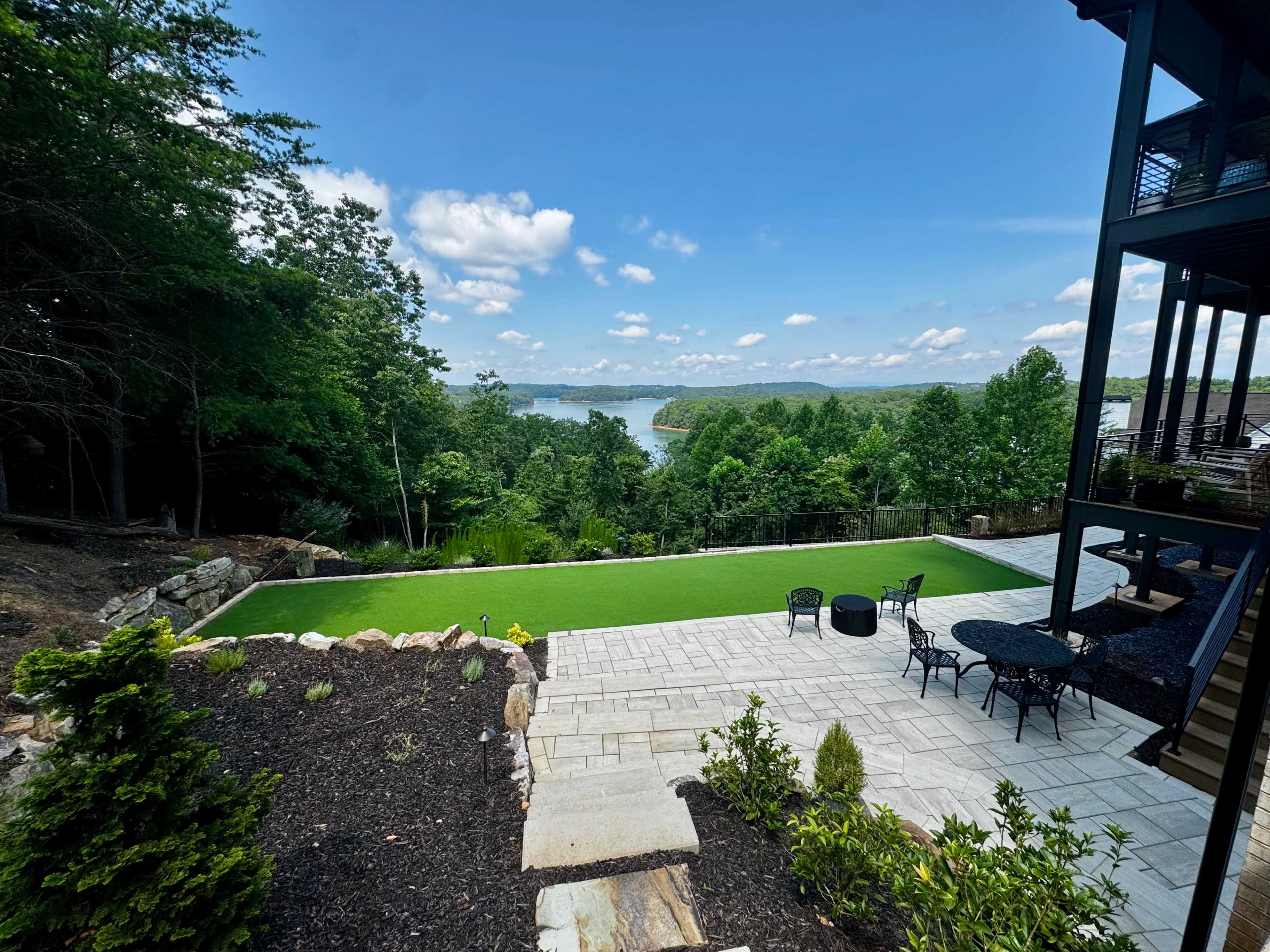 The image shows a landscaped backyard with a manicured lawn overlooking a lake surrounded by trees and a stone patio with outdoor furniture.