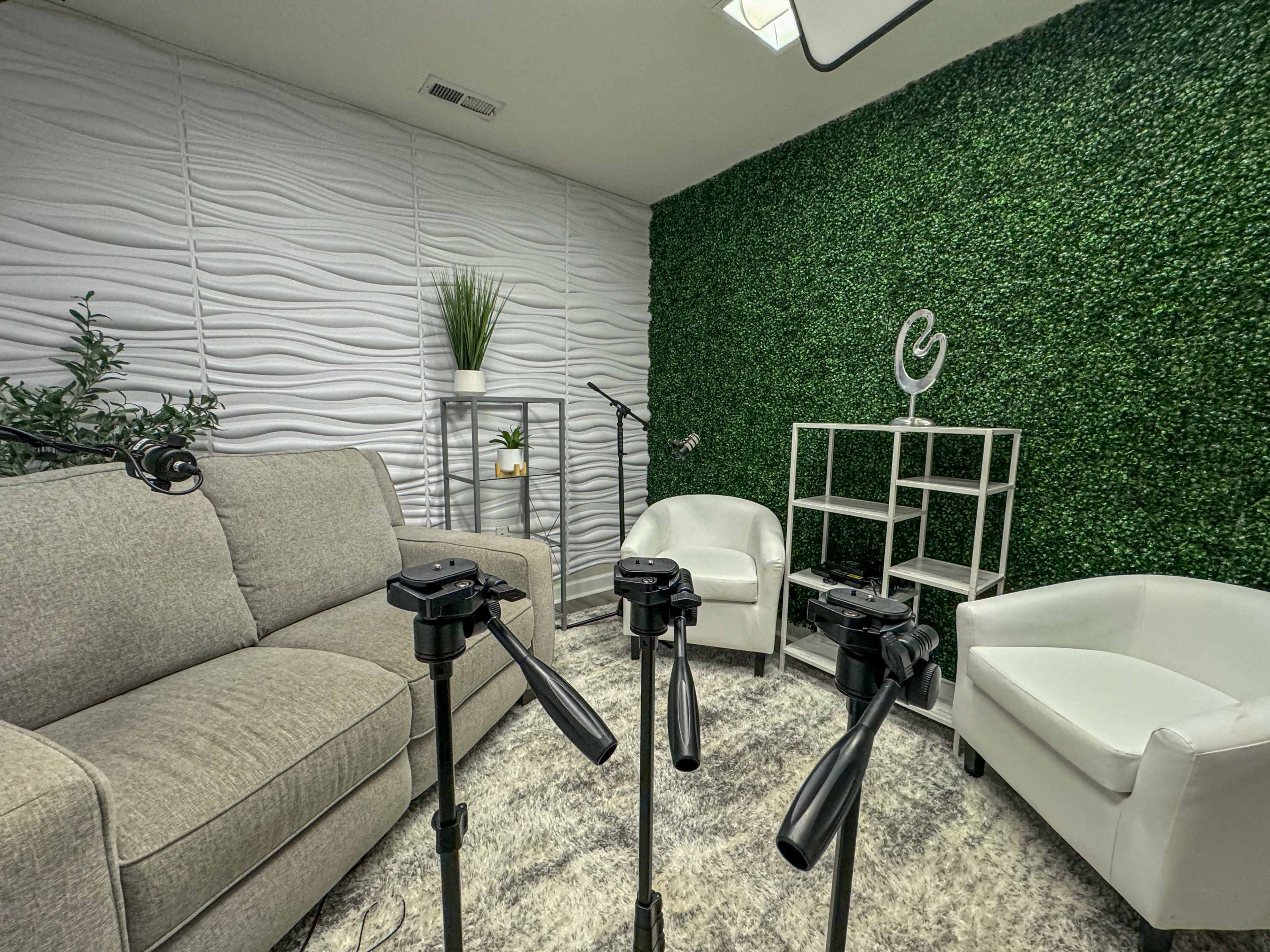 The image shows a small studio space featuring a gray couch, two white chairs, and green wall decor, with microphone stands positioned in front.