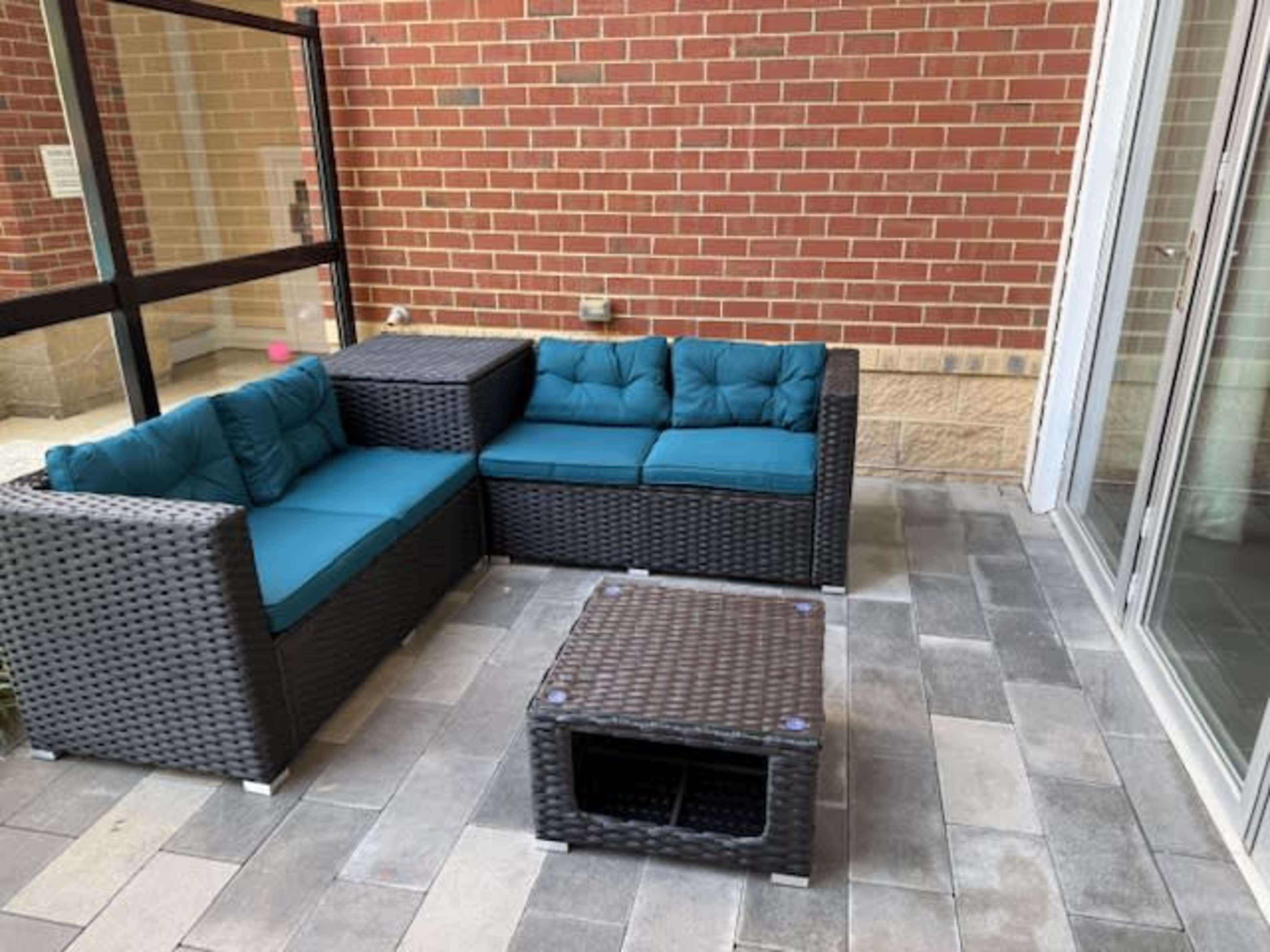 The image shows a patio area with a woven section sofa in dark tones and teal cushions, along with a matching coffee table, against a backdrop of a brick wall.