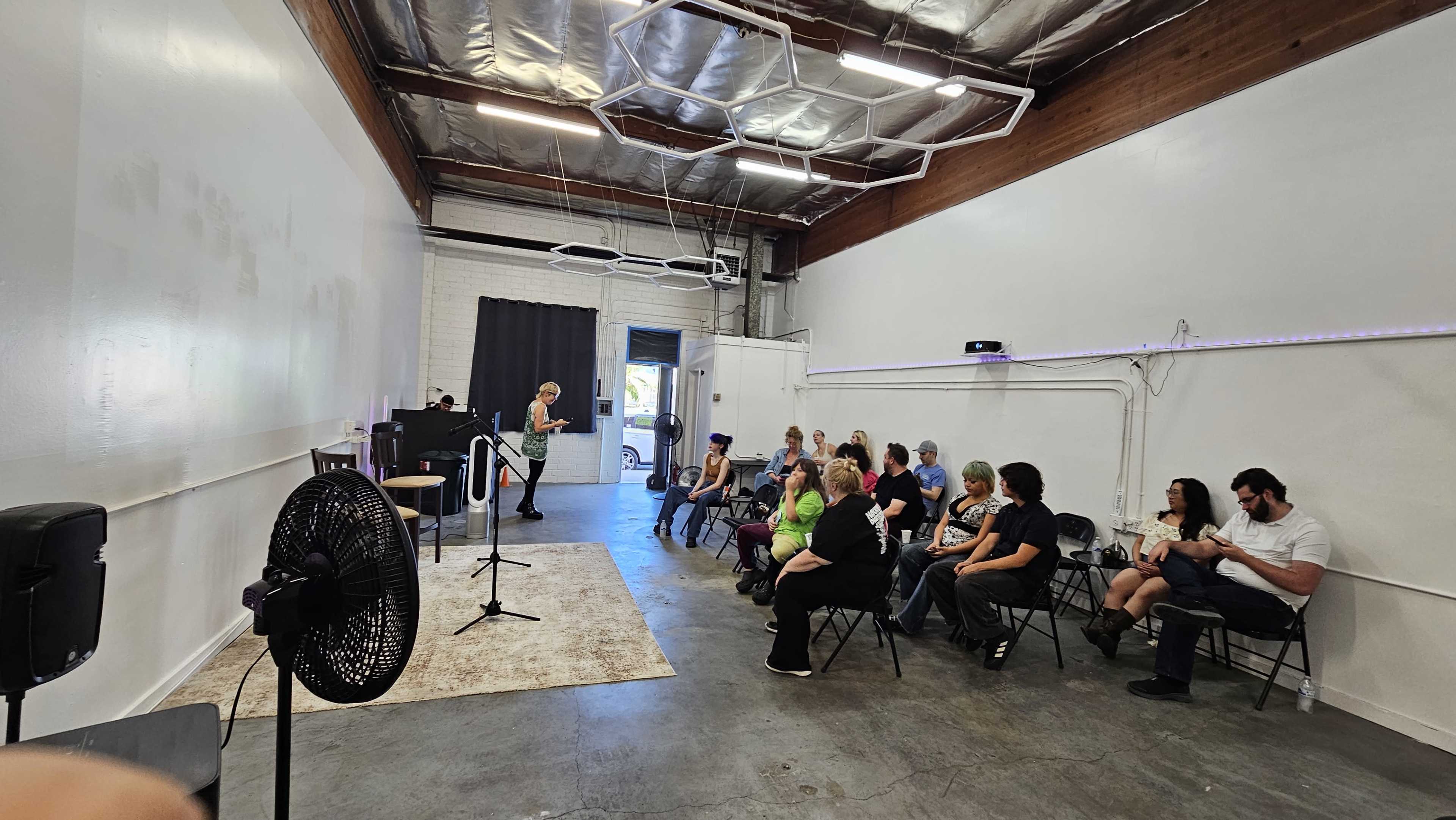 A group of people sits on folding chairs inside a spacious, well-lit room with a stage and musical equipment.