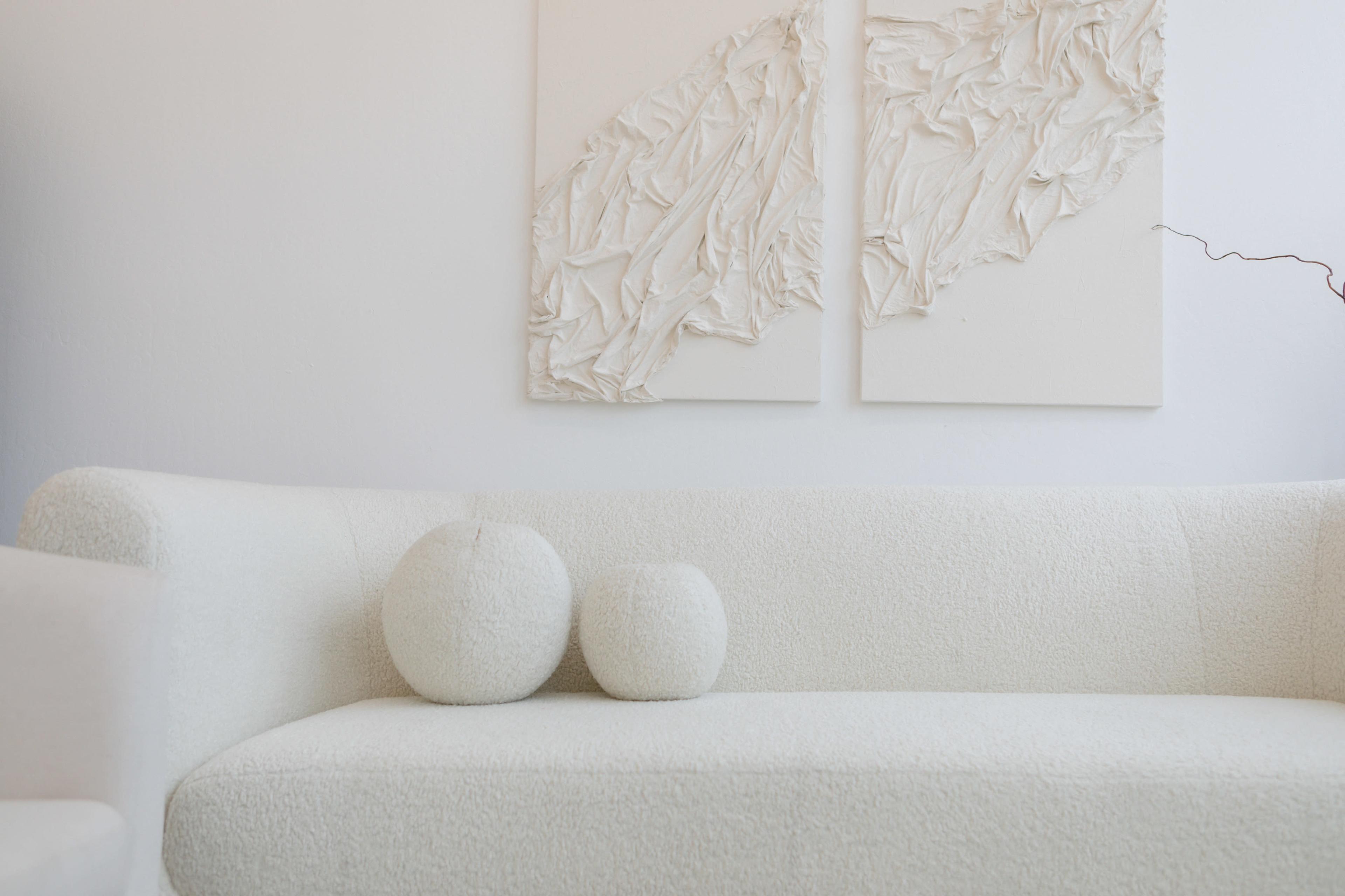 A white, textured sofa with two round cushions is positioned against a wall featuring two abstract, white art pieces.