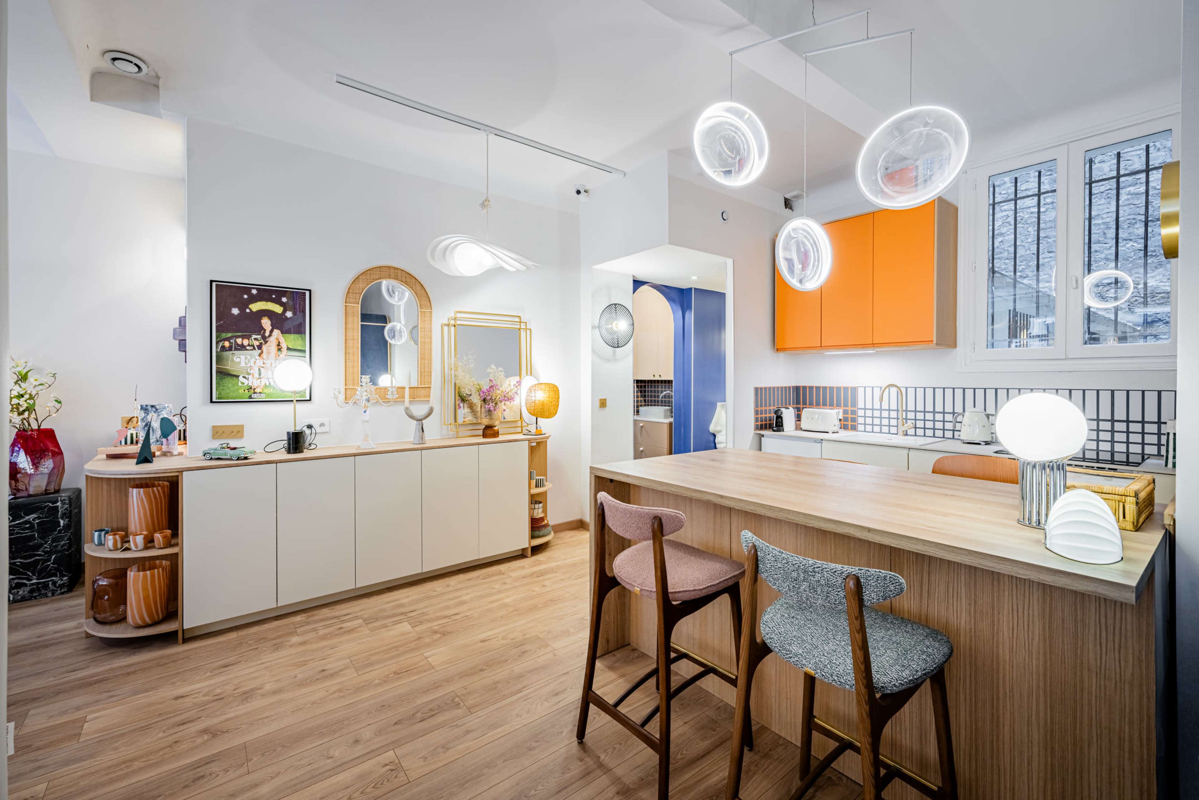 The image shows a modern kitchen with an island, featuring orange cabinets, stylish light fixtures, and a mix of wooden and light-colored materials.