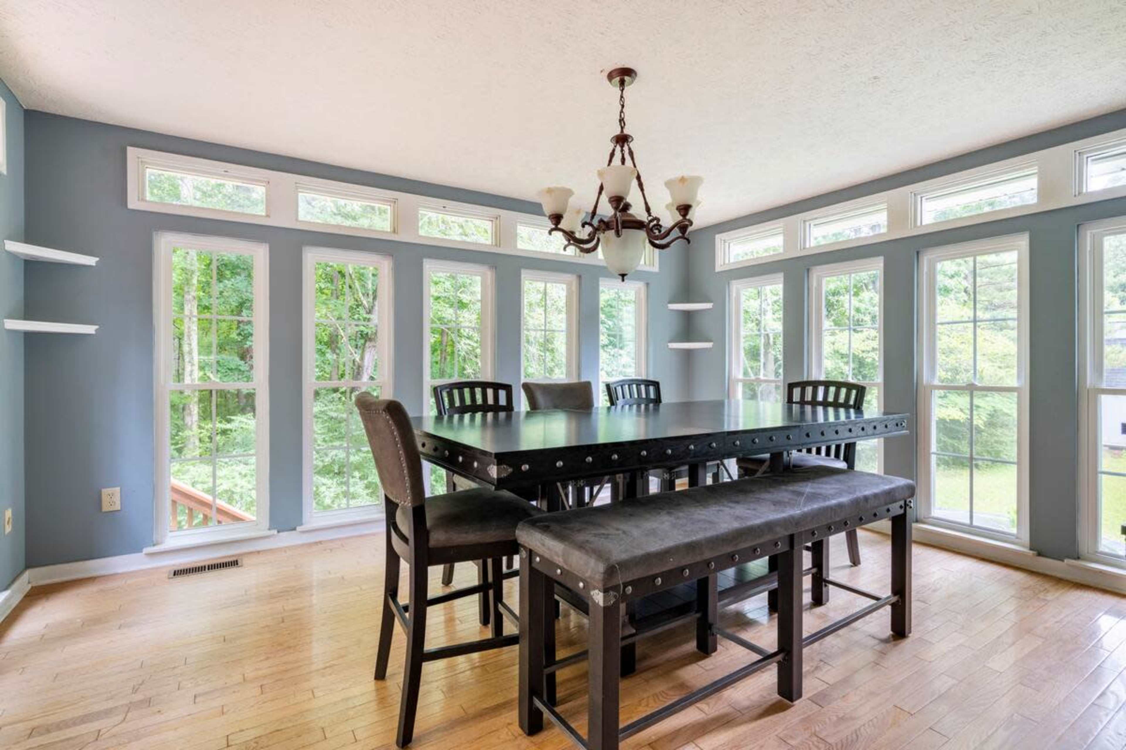 A dining area features a large table and bench with a chandelier, surrounded by tall windows that provide a view of the outdoors.