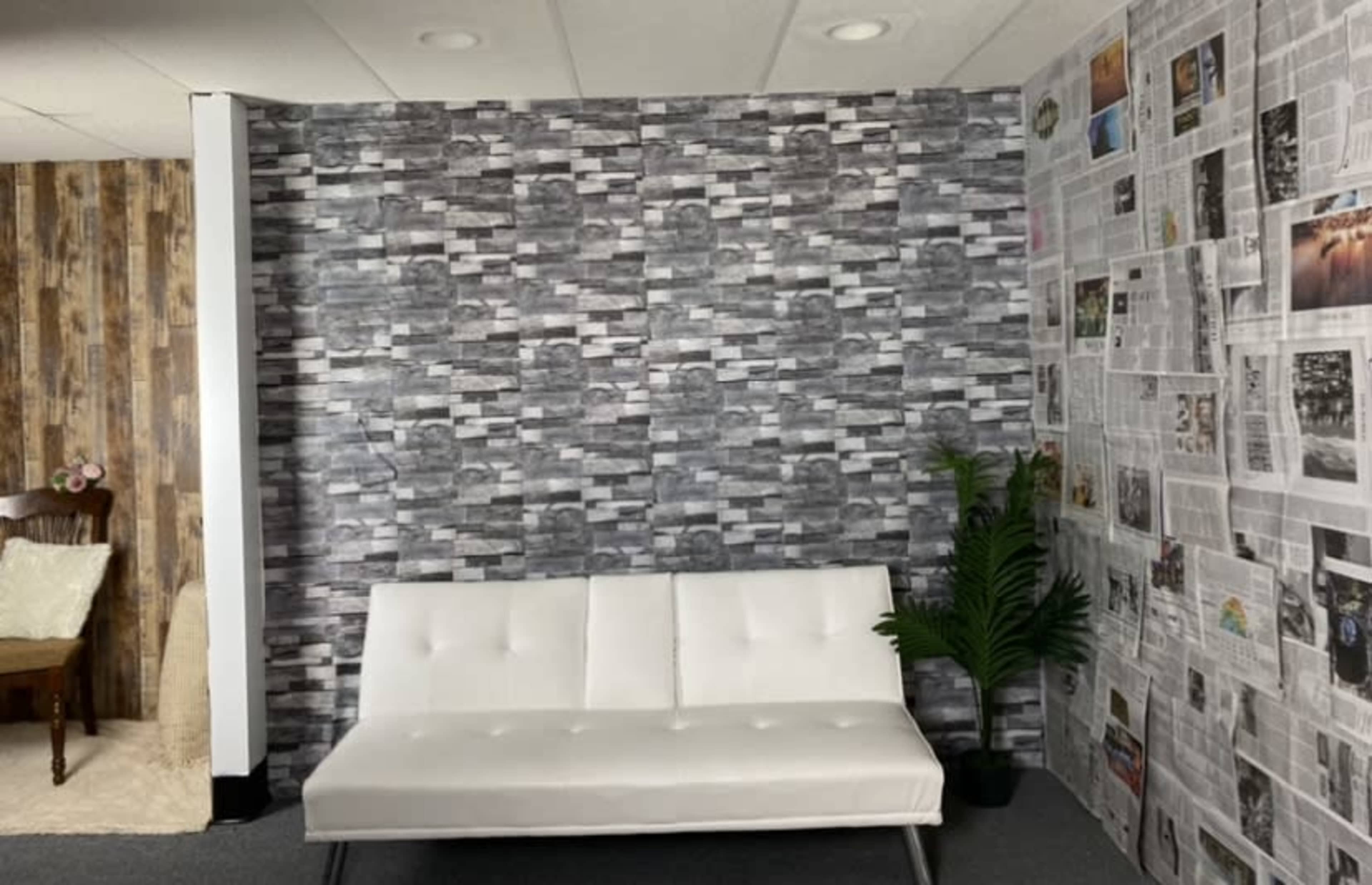 A white sofa is positioned against a gray textured wall, with a nearby wall covered in newspaper clippings and a potted plant in the corner.