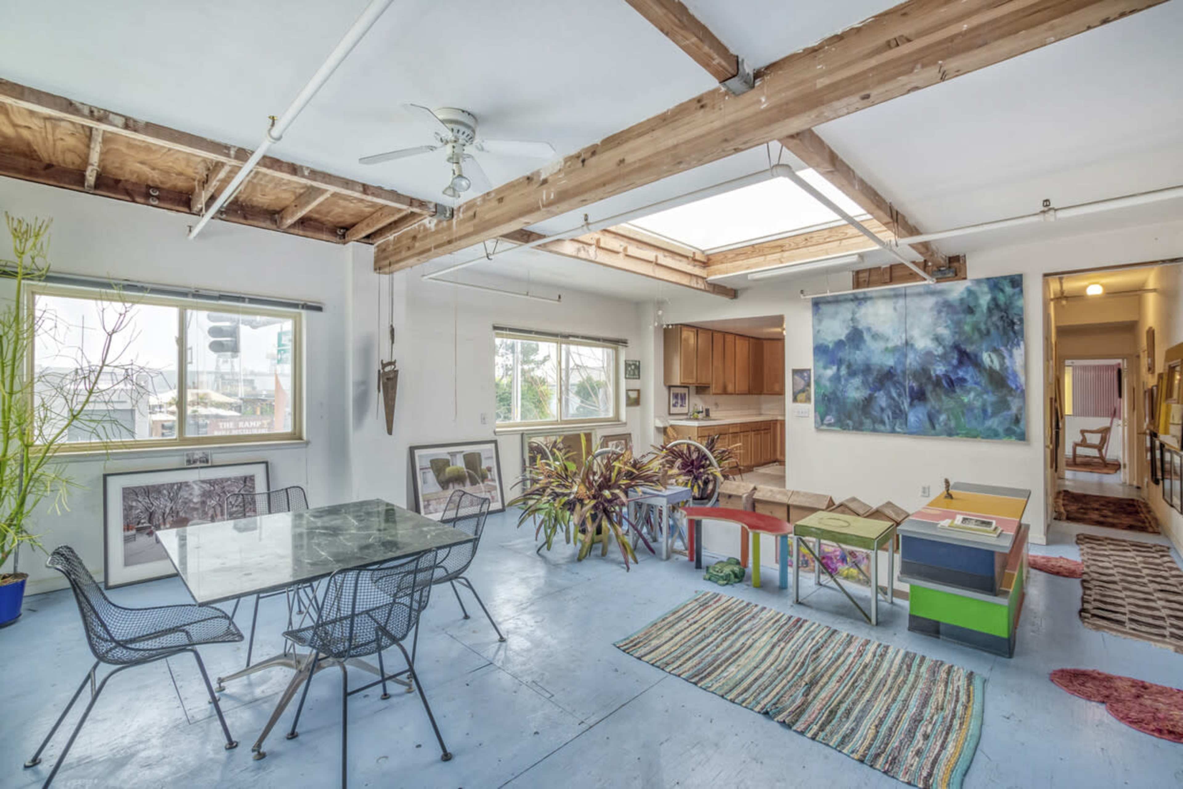 An open-concept room with large windows, a dining table, colorful benches, and a hallway leading to a kitchen.