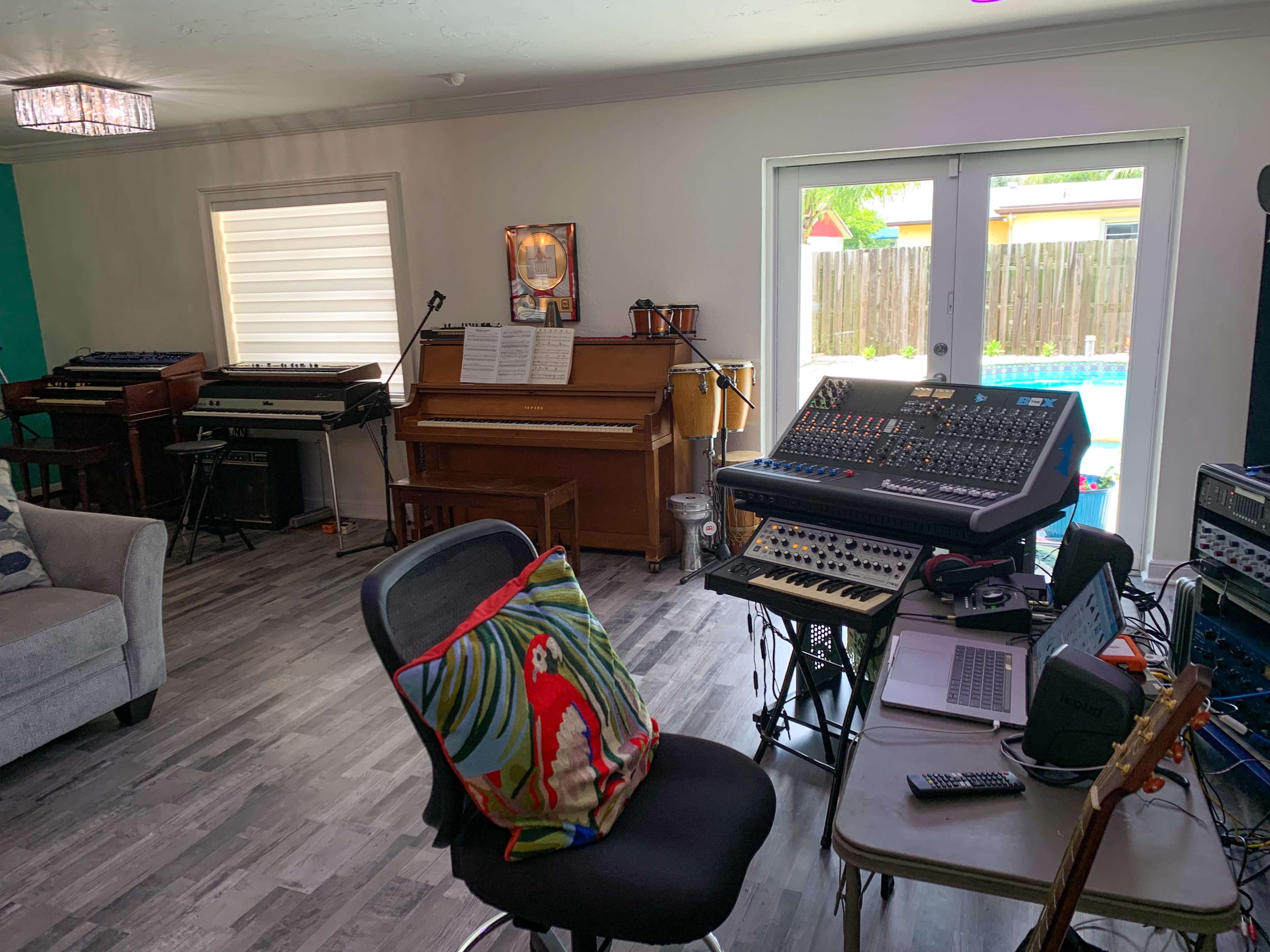 The image shows a home music studio featuring various instruments, a mixing console, a piano, and a view of a pool through sliding glass doors.