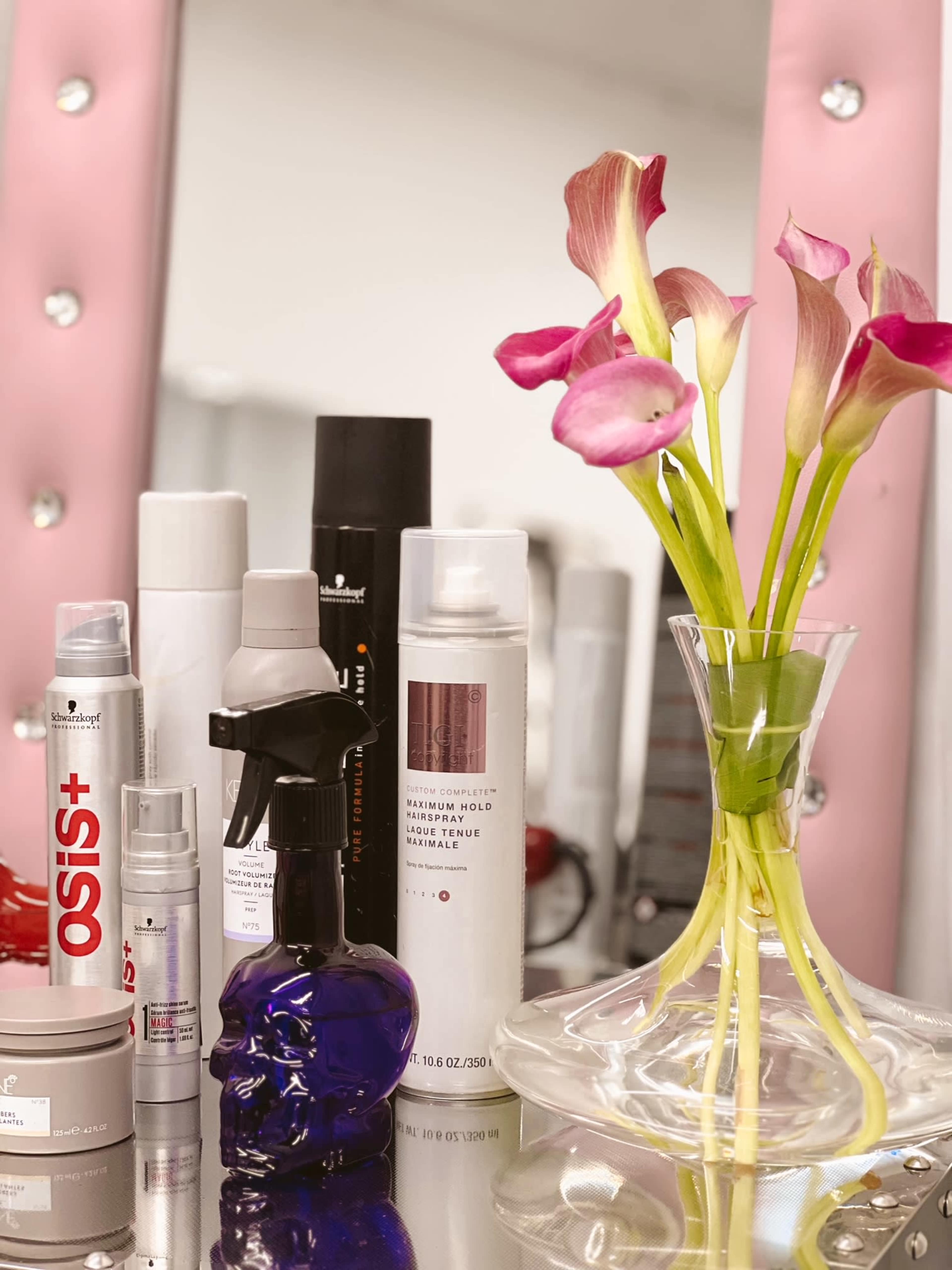 A vanity mirror with various hair and beauty products arranged alongside a vase of pink calla lilies.