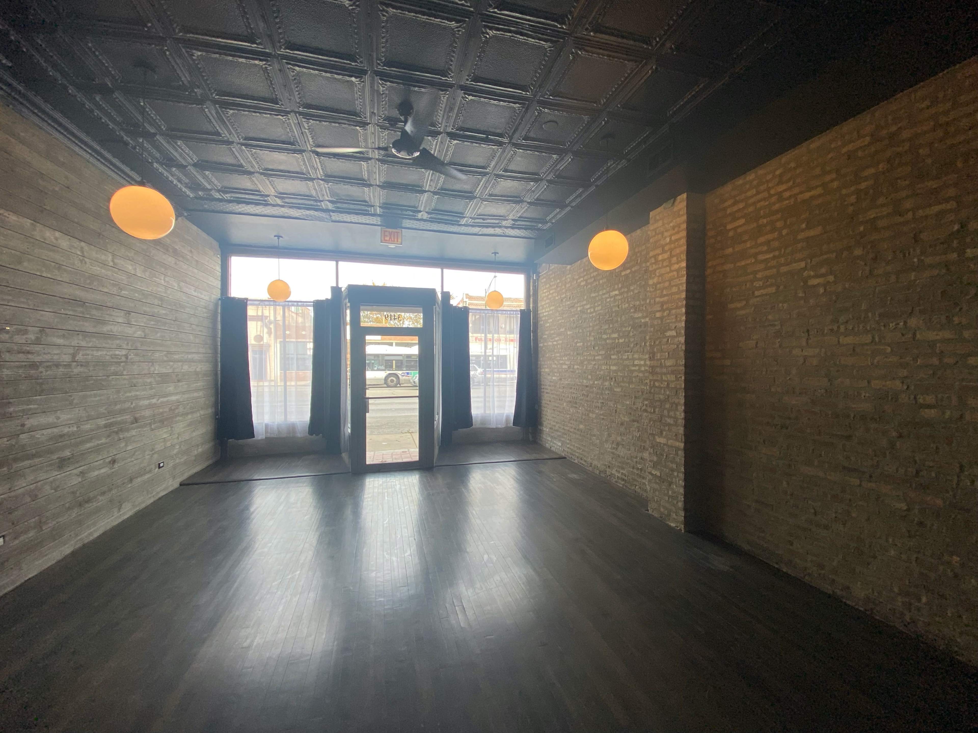 The interior of a vacant commercial space features exposed brick walls, dark wooden flooring, and large windows with hanging light fixtures.