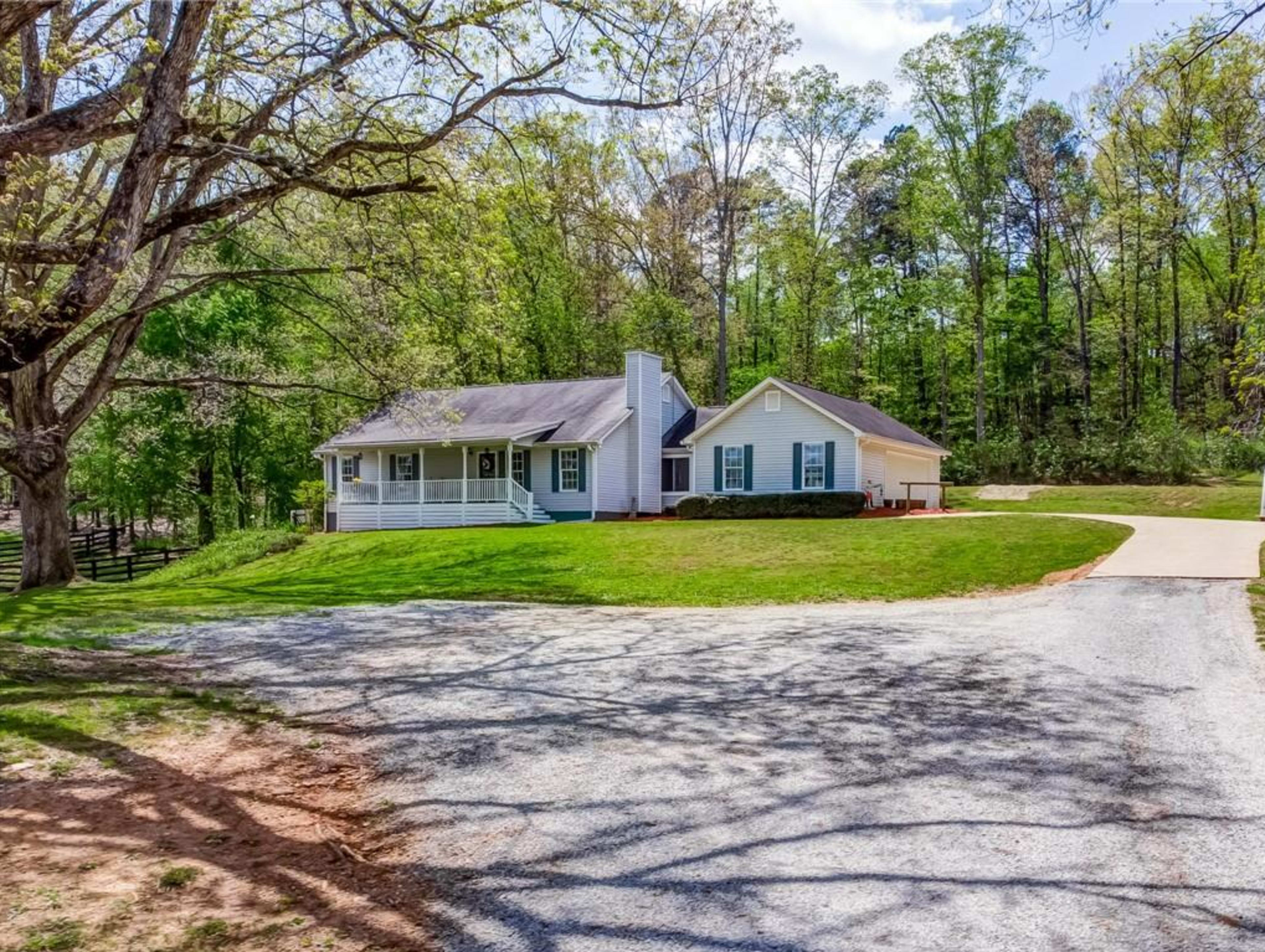A single-story house with a front porch is situated at the end of a gravel driveway, surrounded by trees.