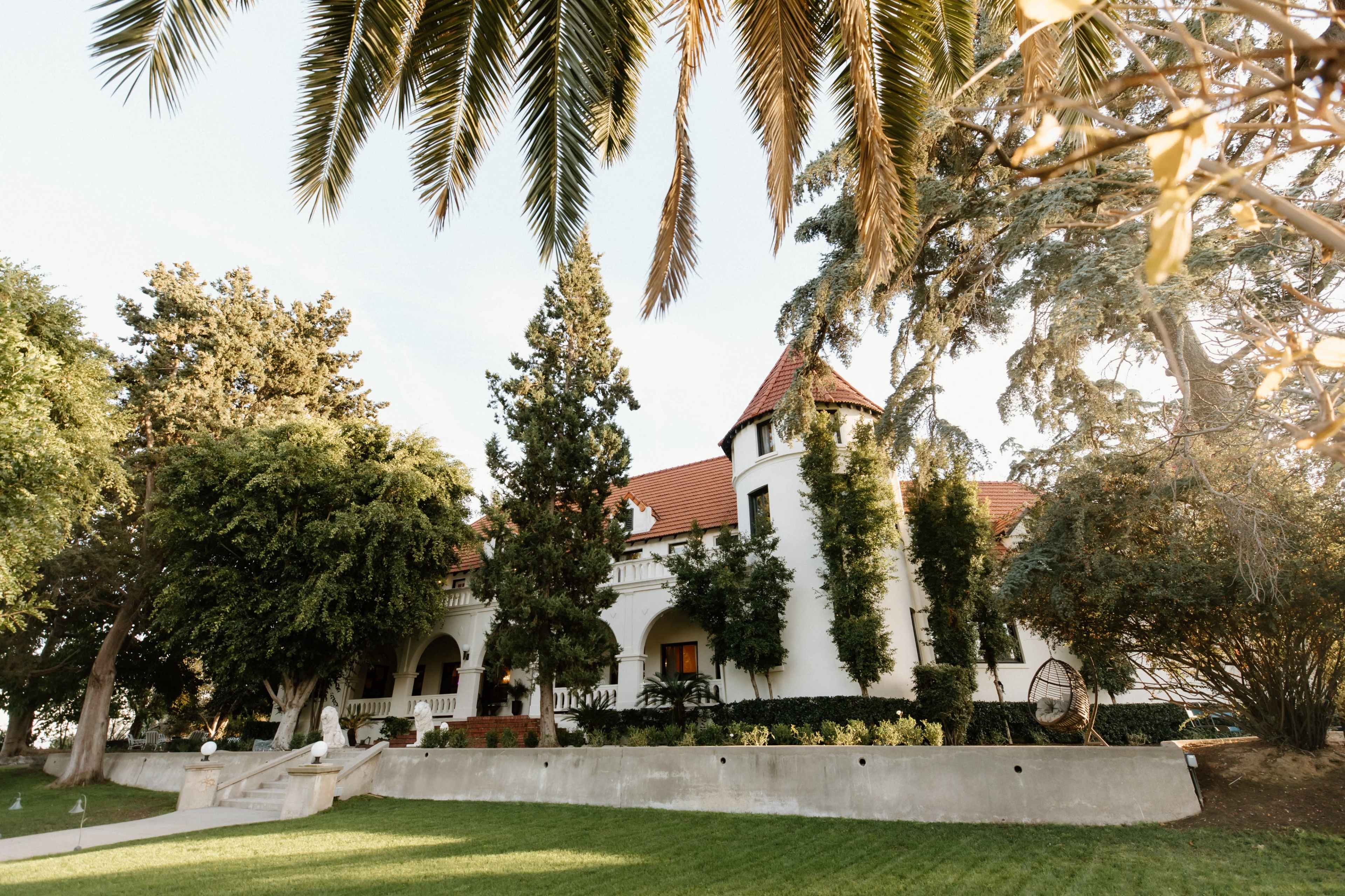 A large, white mansion with a red-tiled roof is surrounded by lush greenery and palm trees.