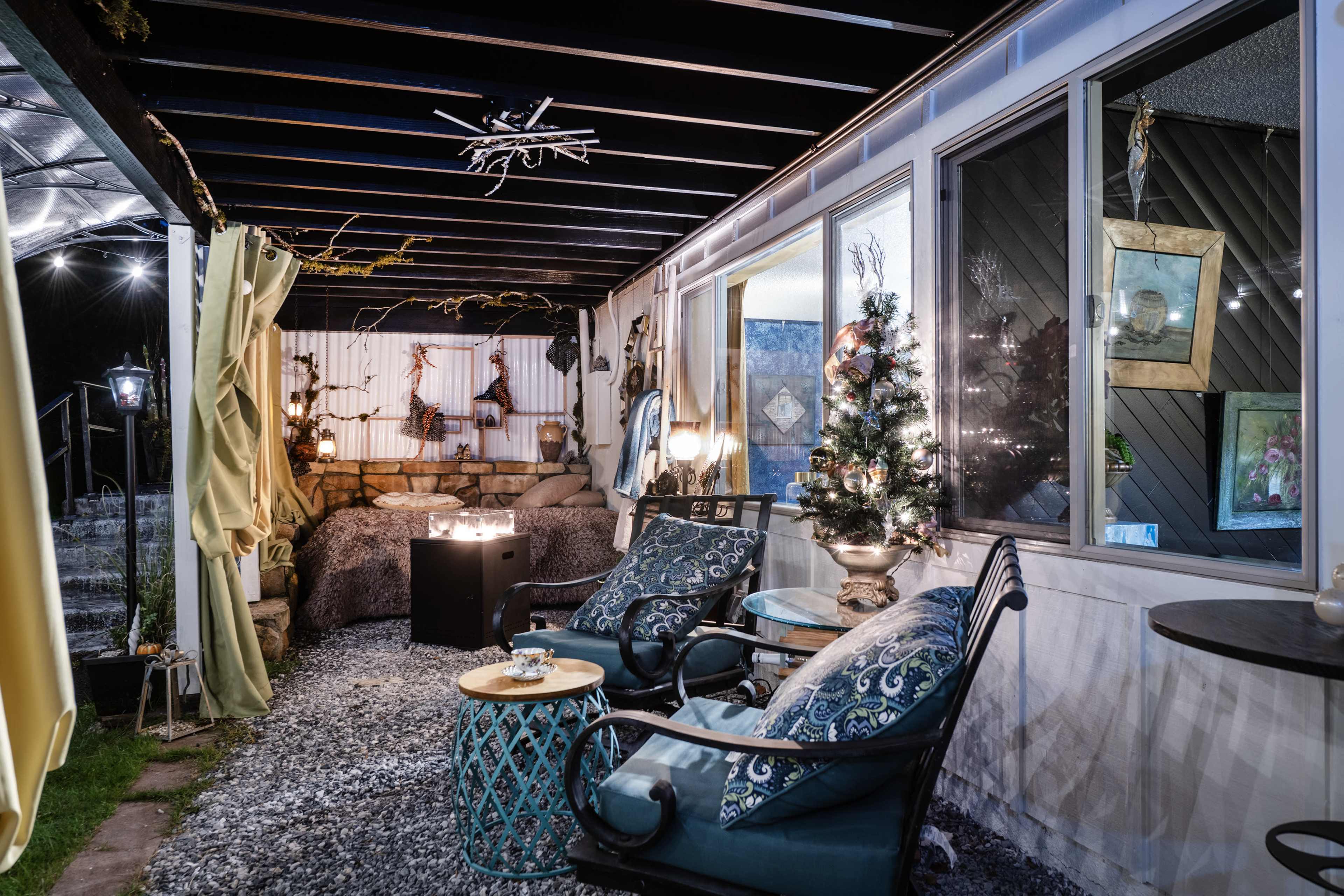 A cozy outdoor seating area features two chairs with decorative cushions, a small table, and various plants, illuminated by string lights and a small tree.