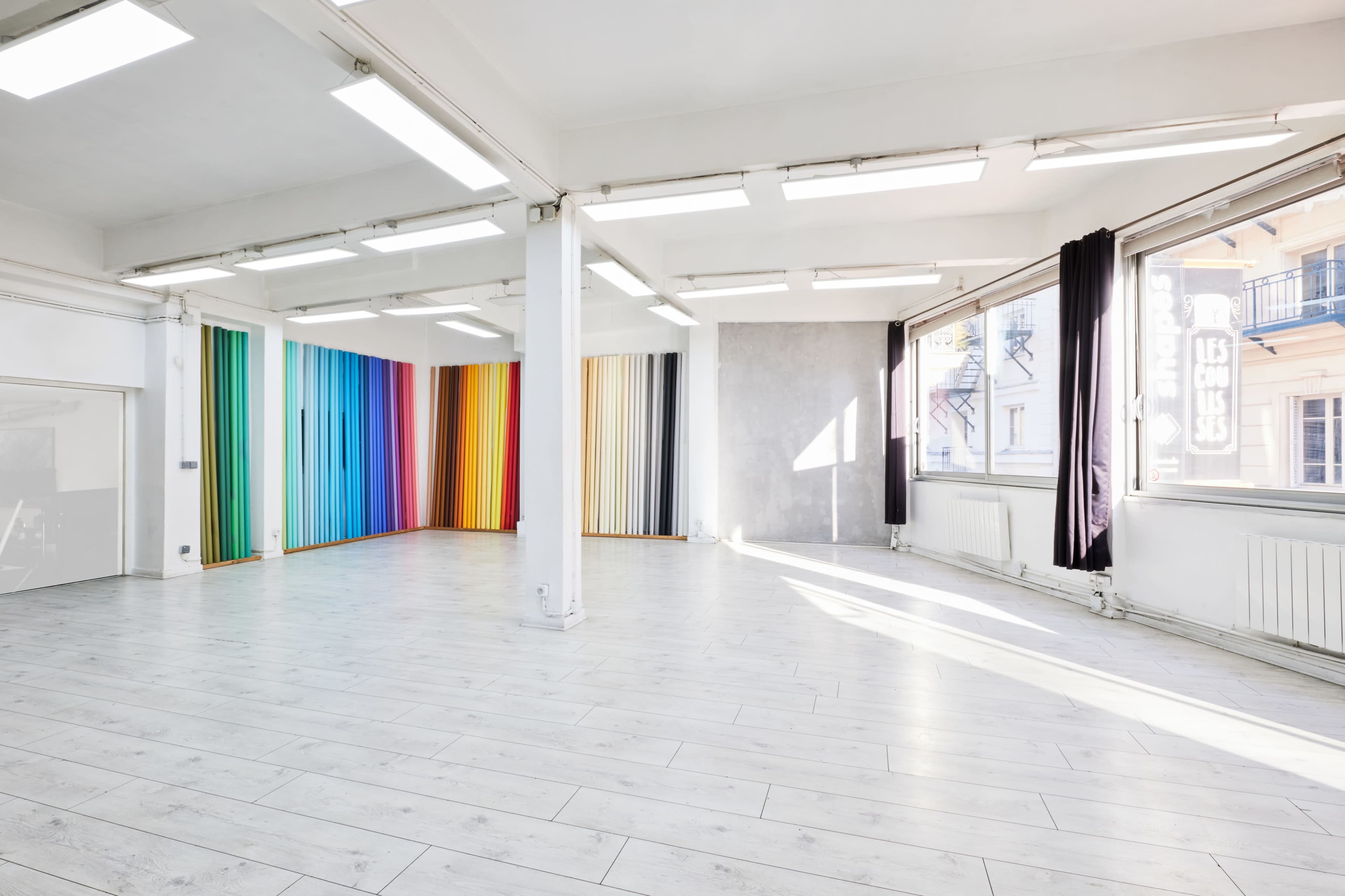 The image shows an empty studio space with colorful fabric panels lining one wall and large windows allowing natural light to enter.