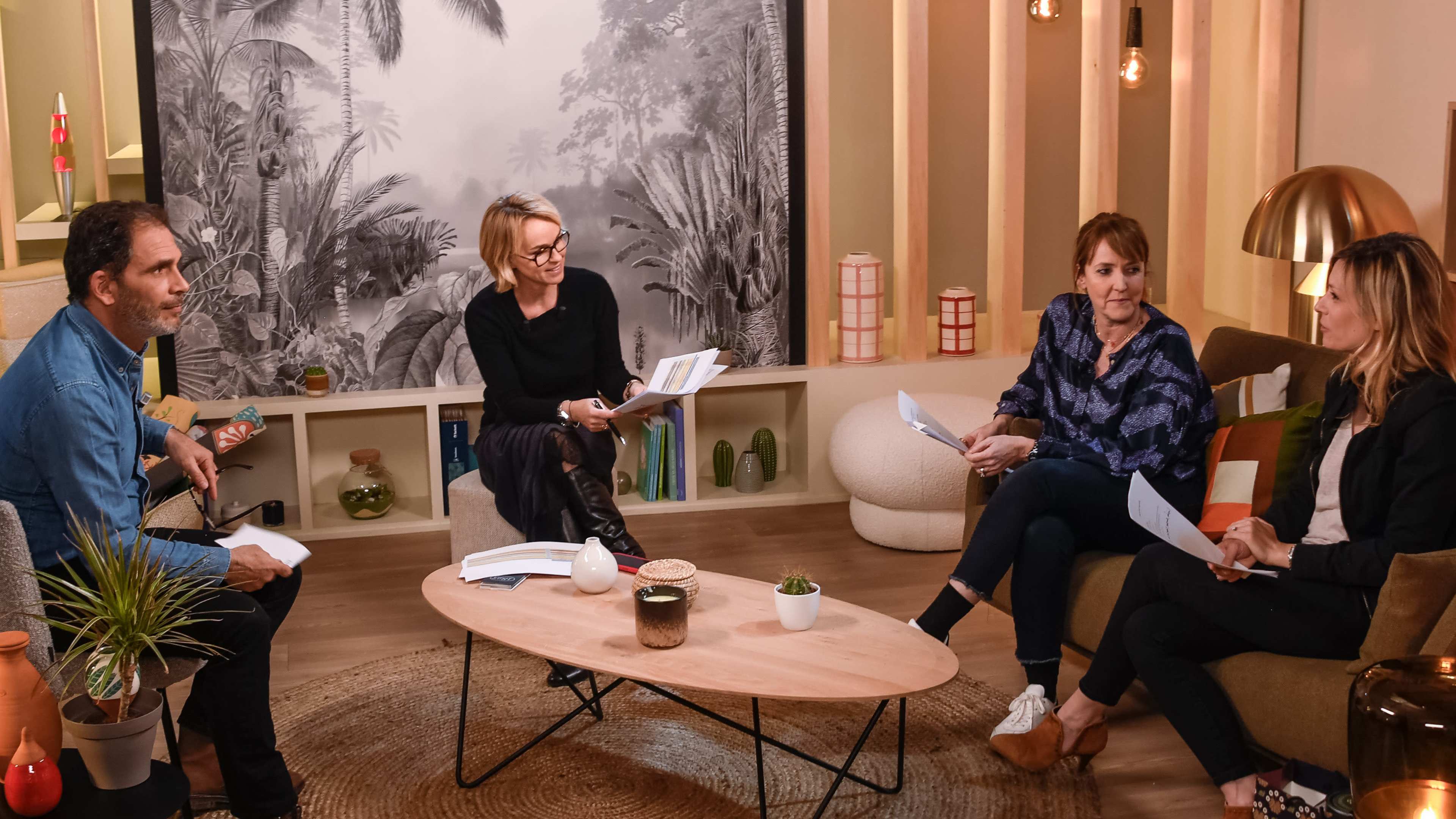 Four people are seated in a softly lit room with a large tropical mural on the wall, engaged in what appears to be a discussion or meeting while holding papers.