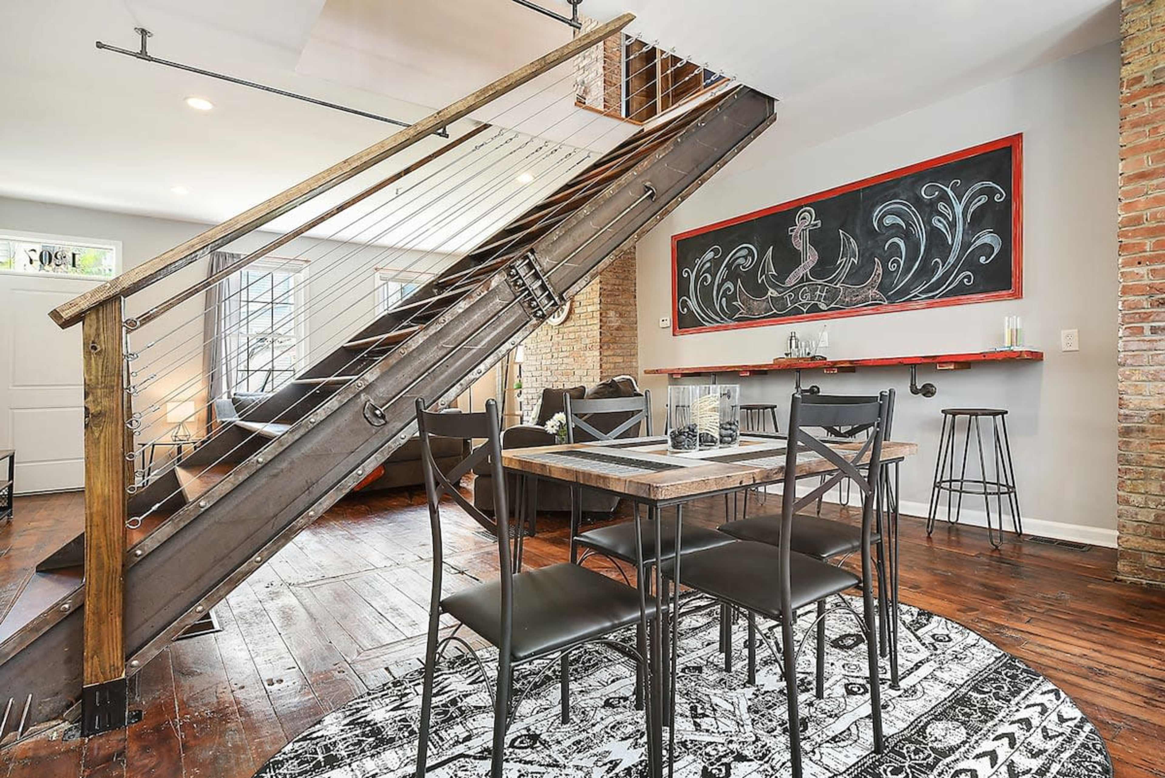 A modern interior featuring a wooden staircase, a dining table with black chairs, and a decorative chalkboard wall in a spacious room with exposed brick.