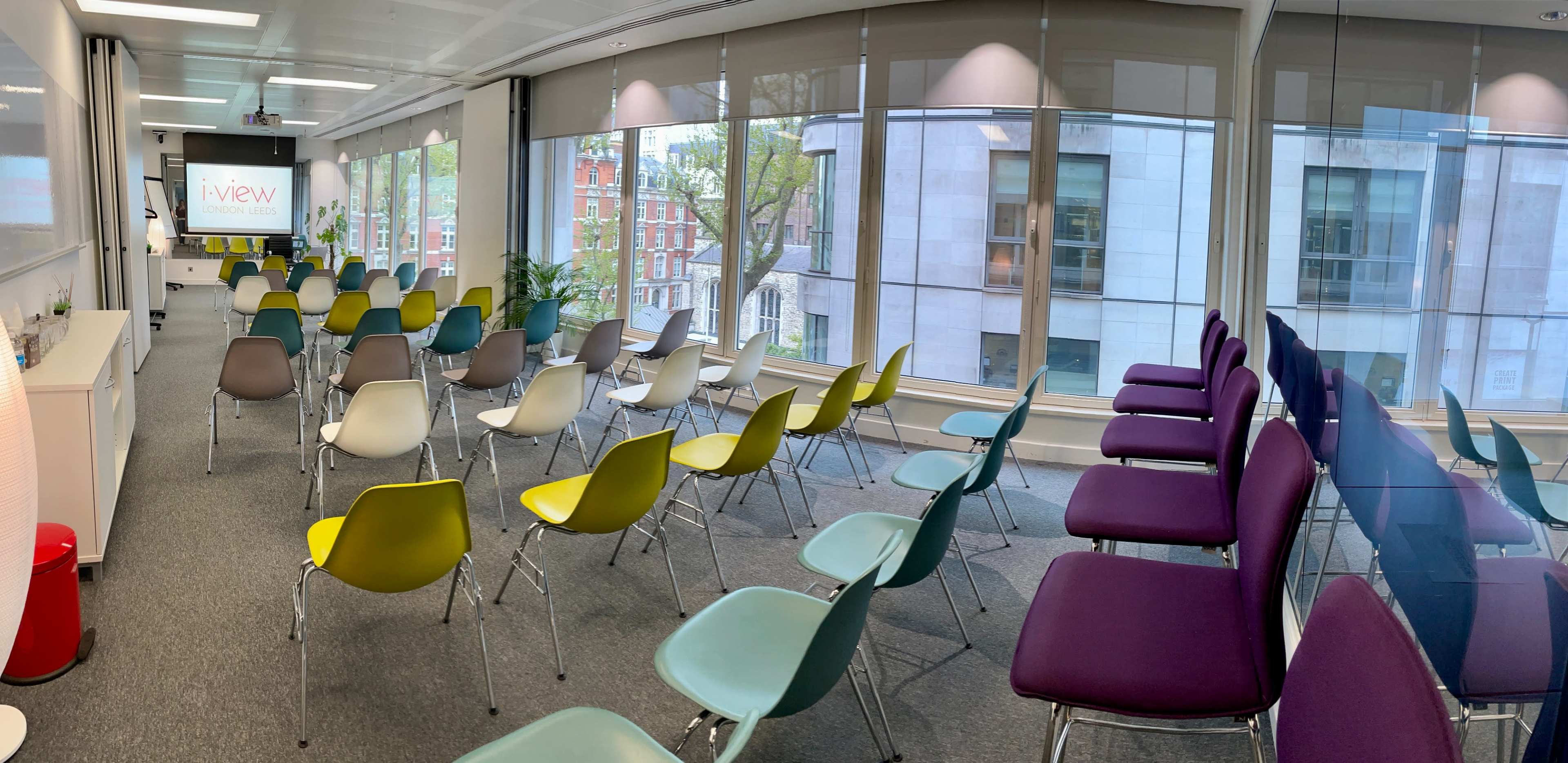 A meeting room with rows of colorful chairs arranged facing a presentation screen and large windows.