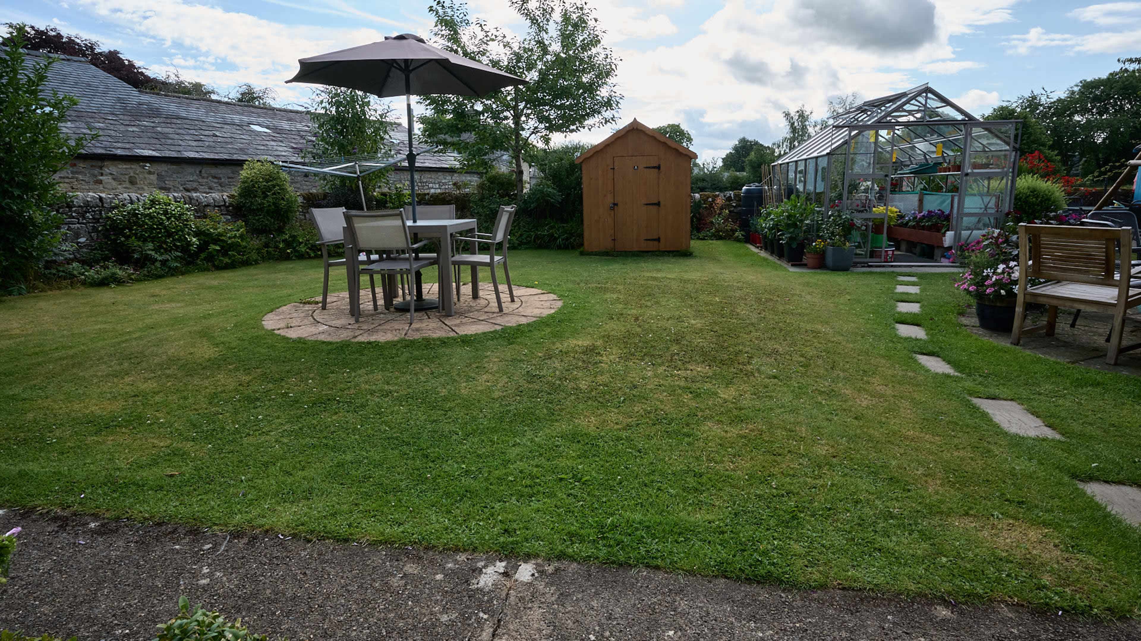 The image shows a well-maintained garden featuring a circular patio with a table and chairs, a wooden shed, and a greenhouse in the background.