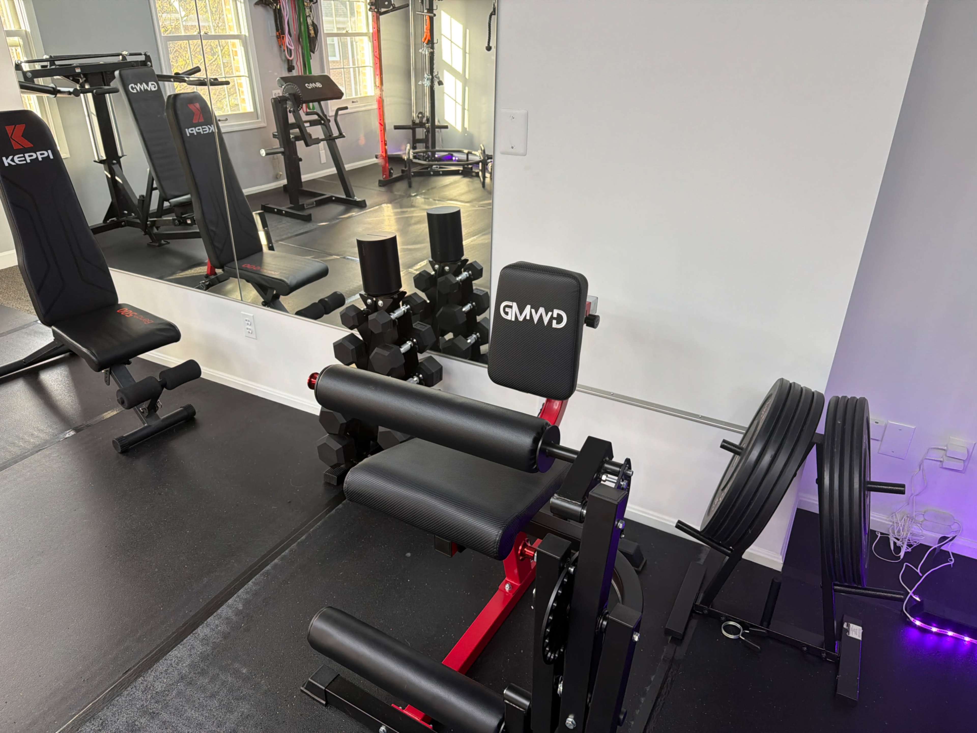 The image shows a home gym setup with a black and red piece of exercise equipment, free weights, and a mirrored wall reflecting additional gym equipment in the background.