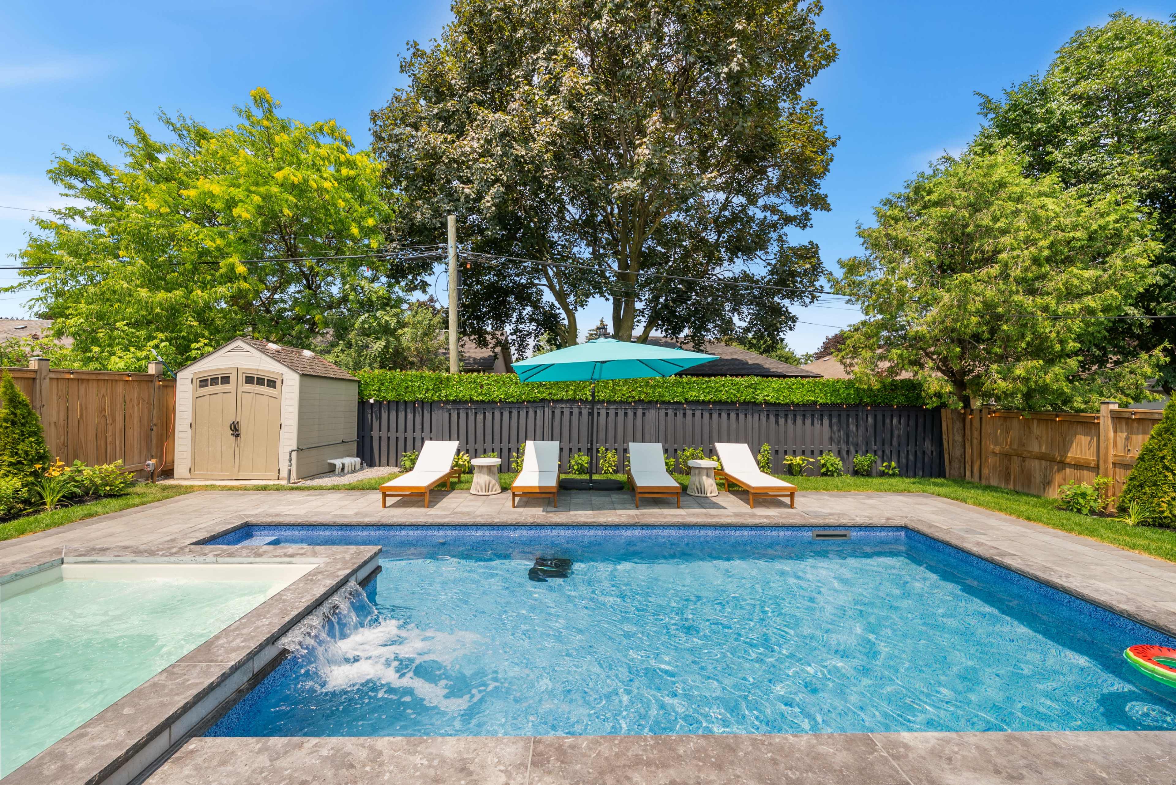 The image shows a backyard pool area featuring a swimming pool with lounge chairs, an umbrella, and a storage shed surrounded by trees and a wooden fence.
