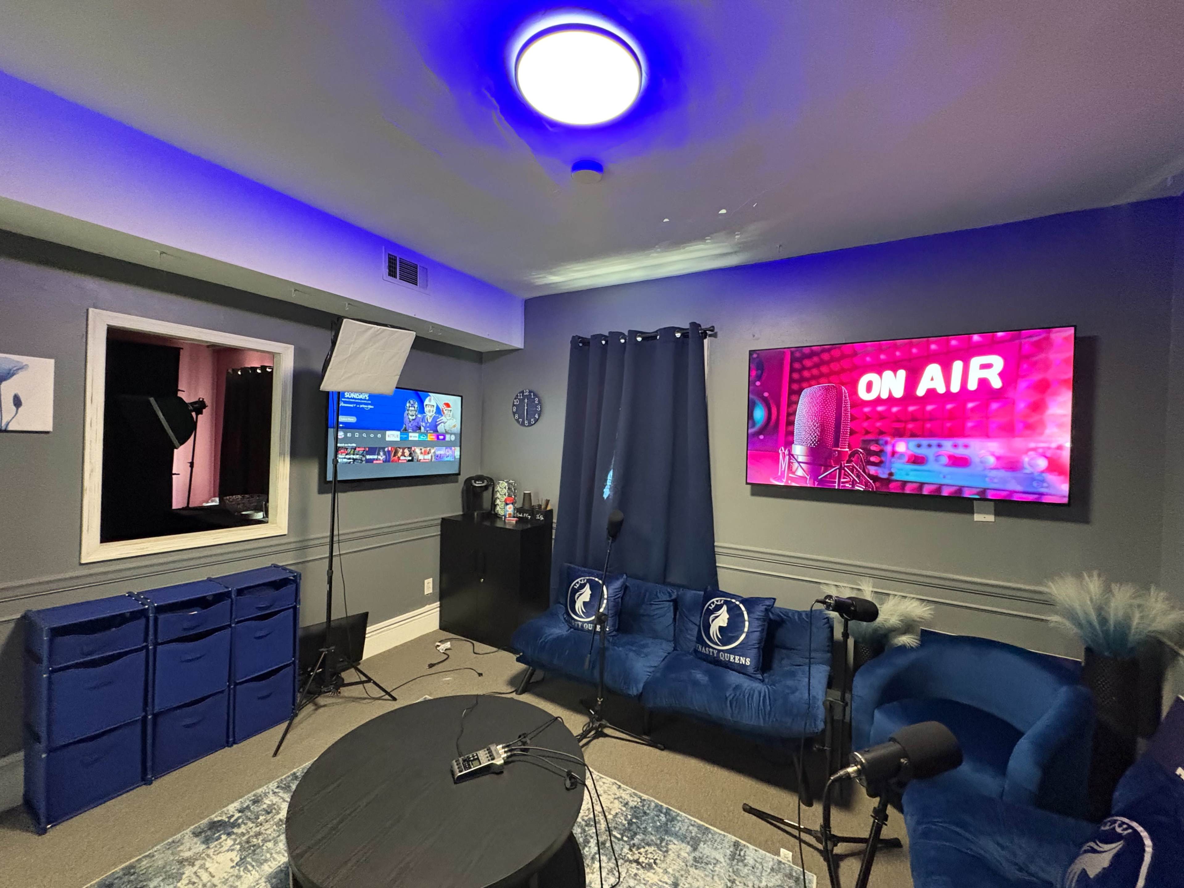 The image depicts a studio room featuring two large screens, a dark blue couch, a table, and a window with a black curtain, all arranged for a media production setup.