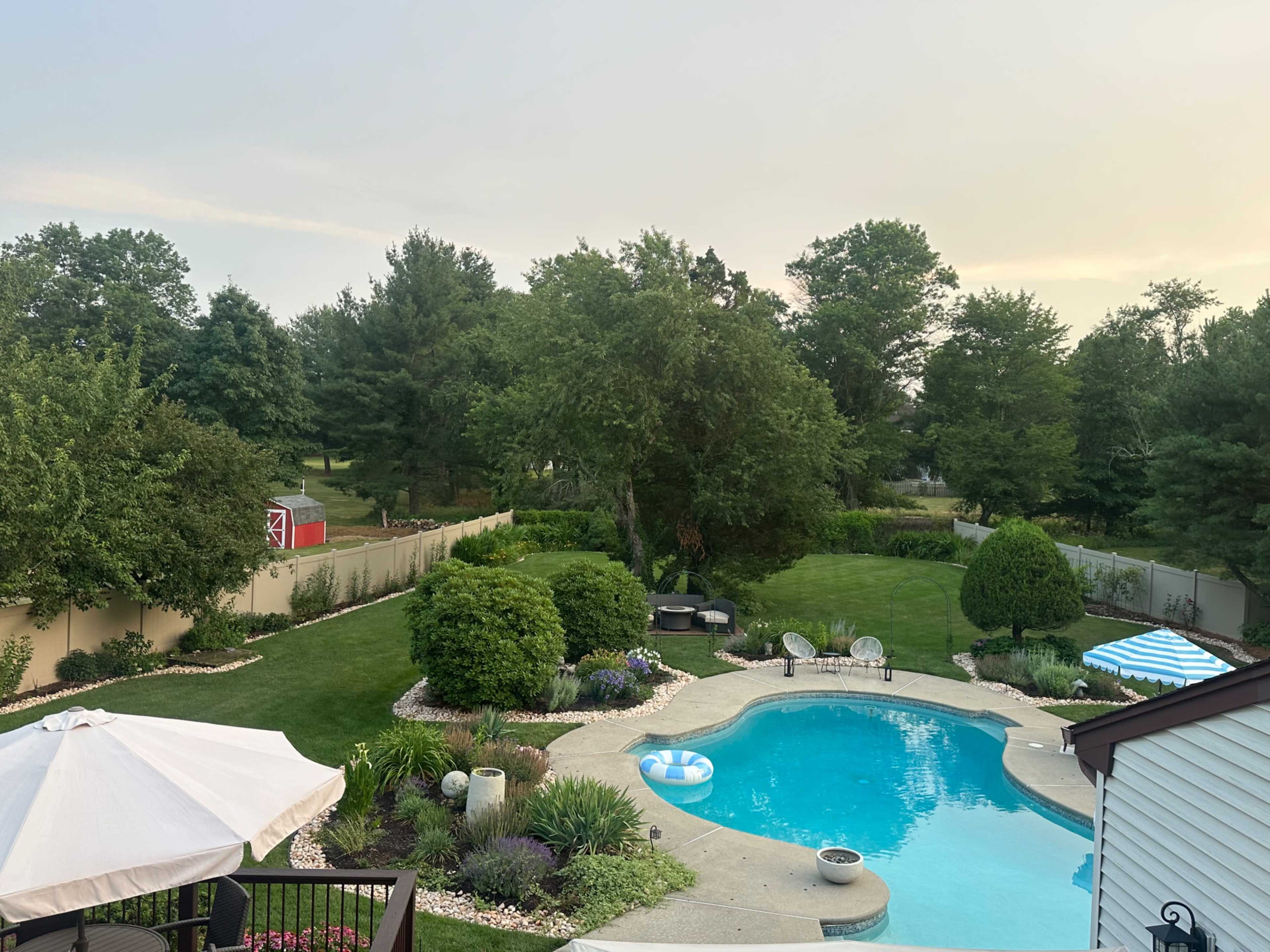 The image shows a residential backyard featuring a swimming pool surrounded by lush greenery, patio furniture, and decorative landscaping.