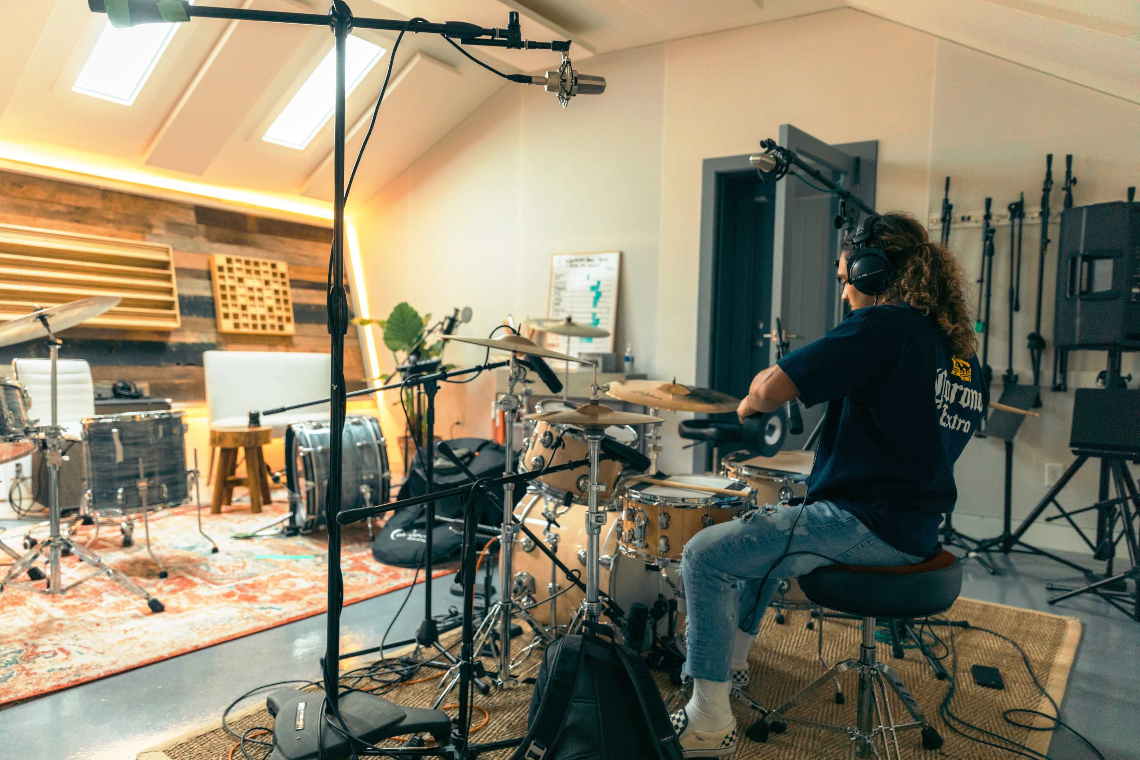 A drummer is seated at a drum kit in a well-lit music studio with various instruments and sound equipment visible.