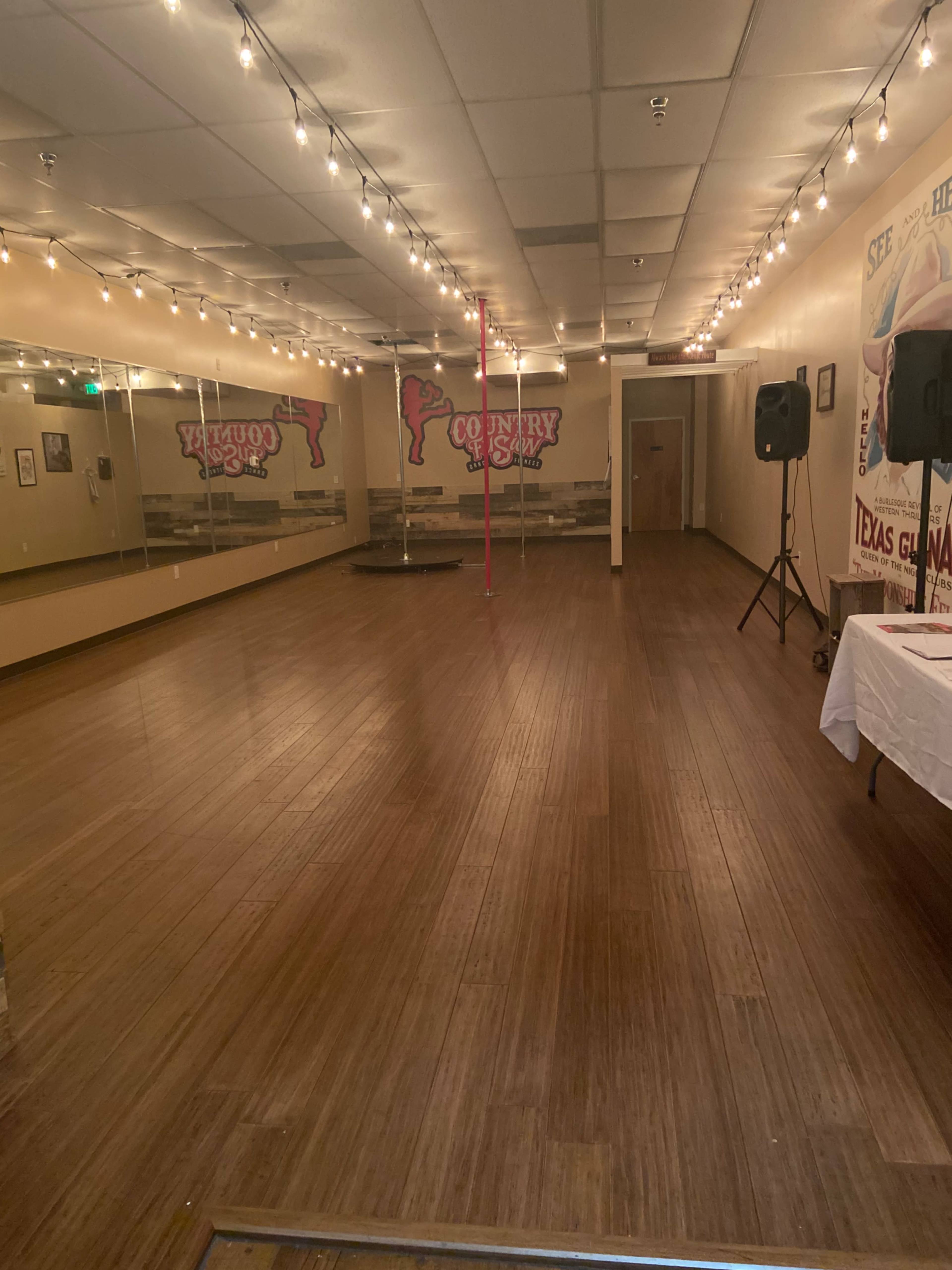 An empty dance studio with wooden floors, mirrored walls, and a pink dance pole in the center, illuminated by overhead string lights.