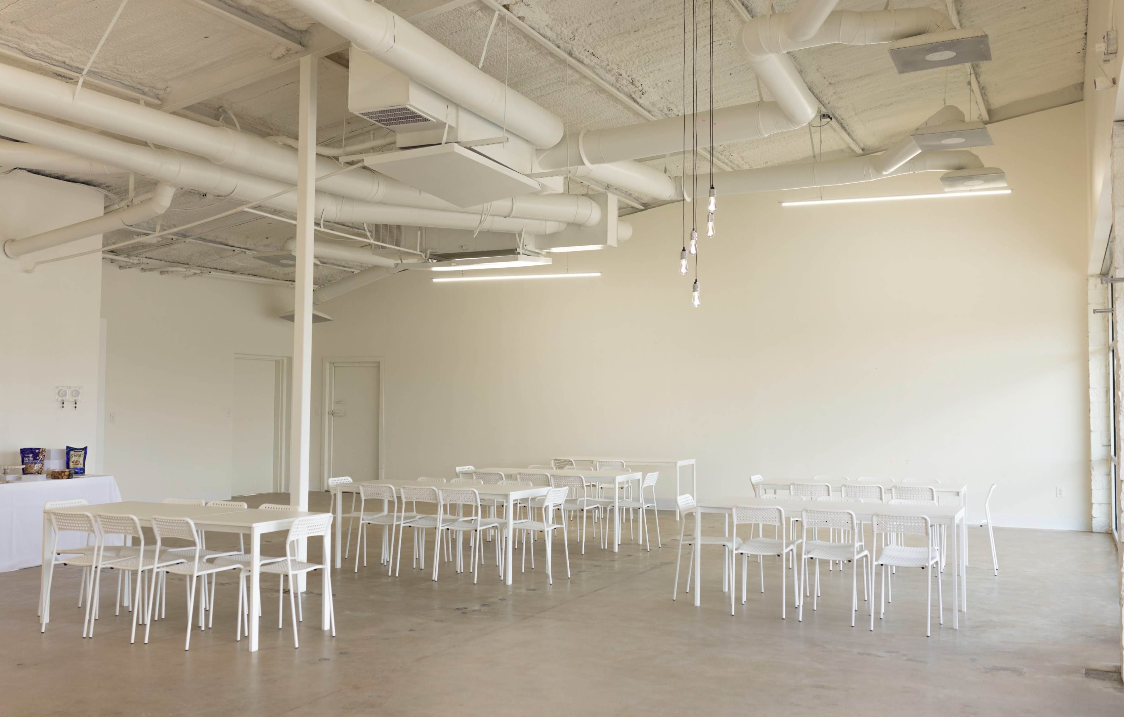 A bright, minimalist room features multiple white tables and chairs arranged on a concrete floor, with exposed ductwork and hanging light fixtures.