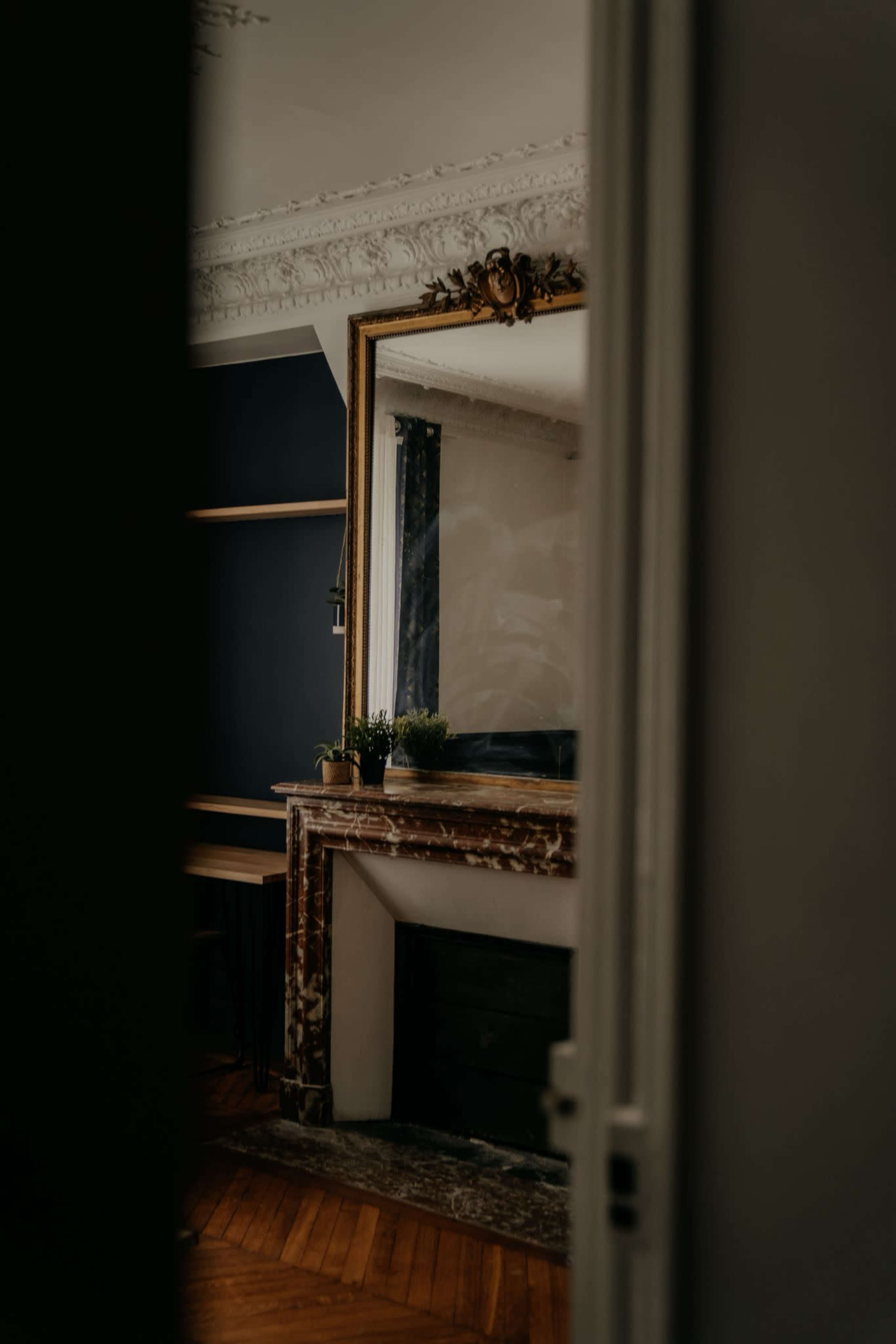 The image shows a glimpse of a room with a marble fireplace and a large ornate mirror framed in gold.