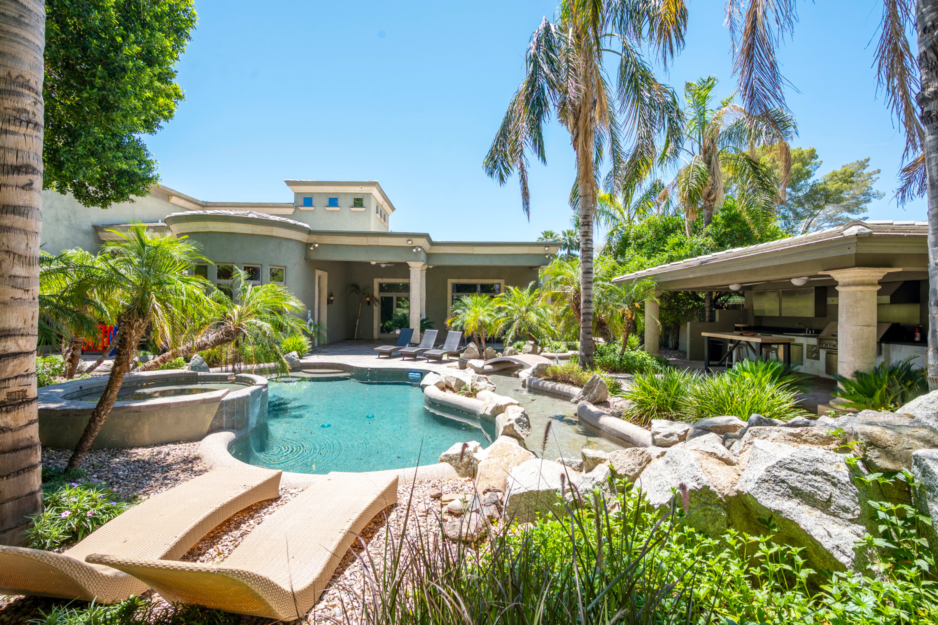 The image shows a luxurious backyard with a swimming pool, surrounded by tropical plants and palm trees, along with a patio area and an outdoor kitchen.
