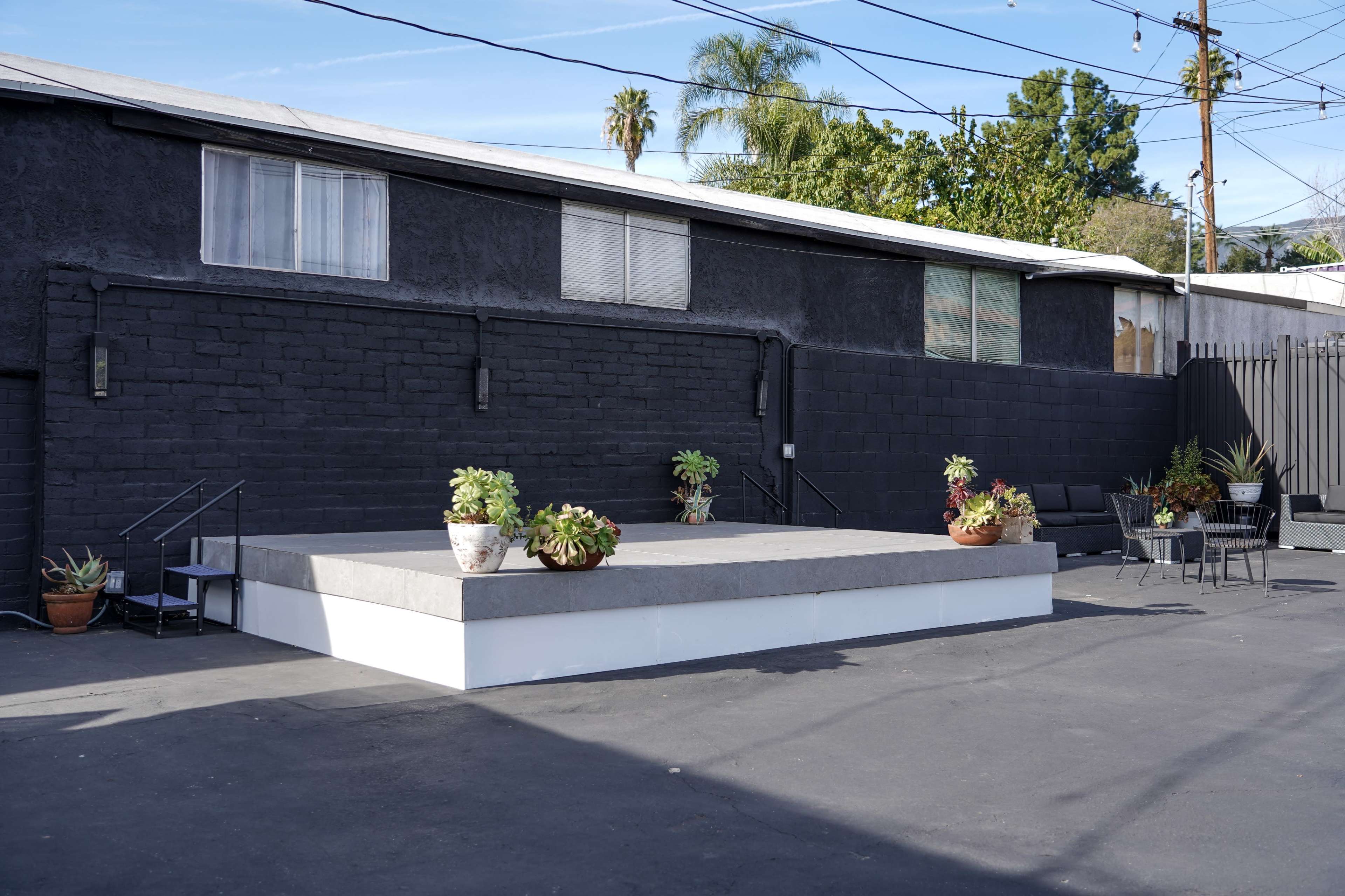 The image shows a small outdoor stage surrounded by potted plants, with a black building and utility poles in the background.