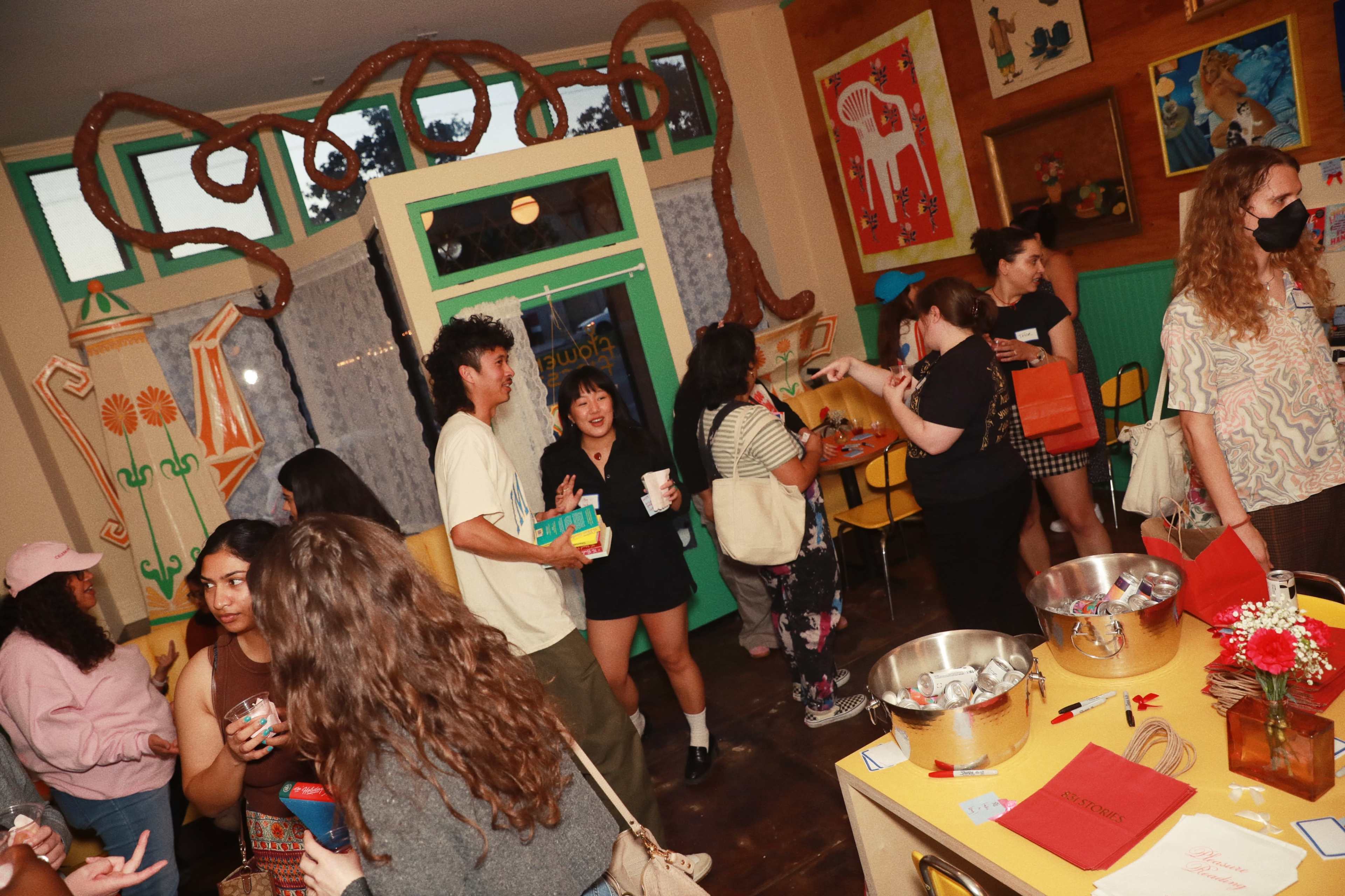 A group of people socialize in a brightly colored room decorated with art and plants while holding drinks and snacks.