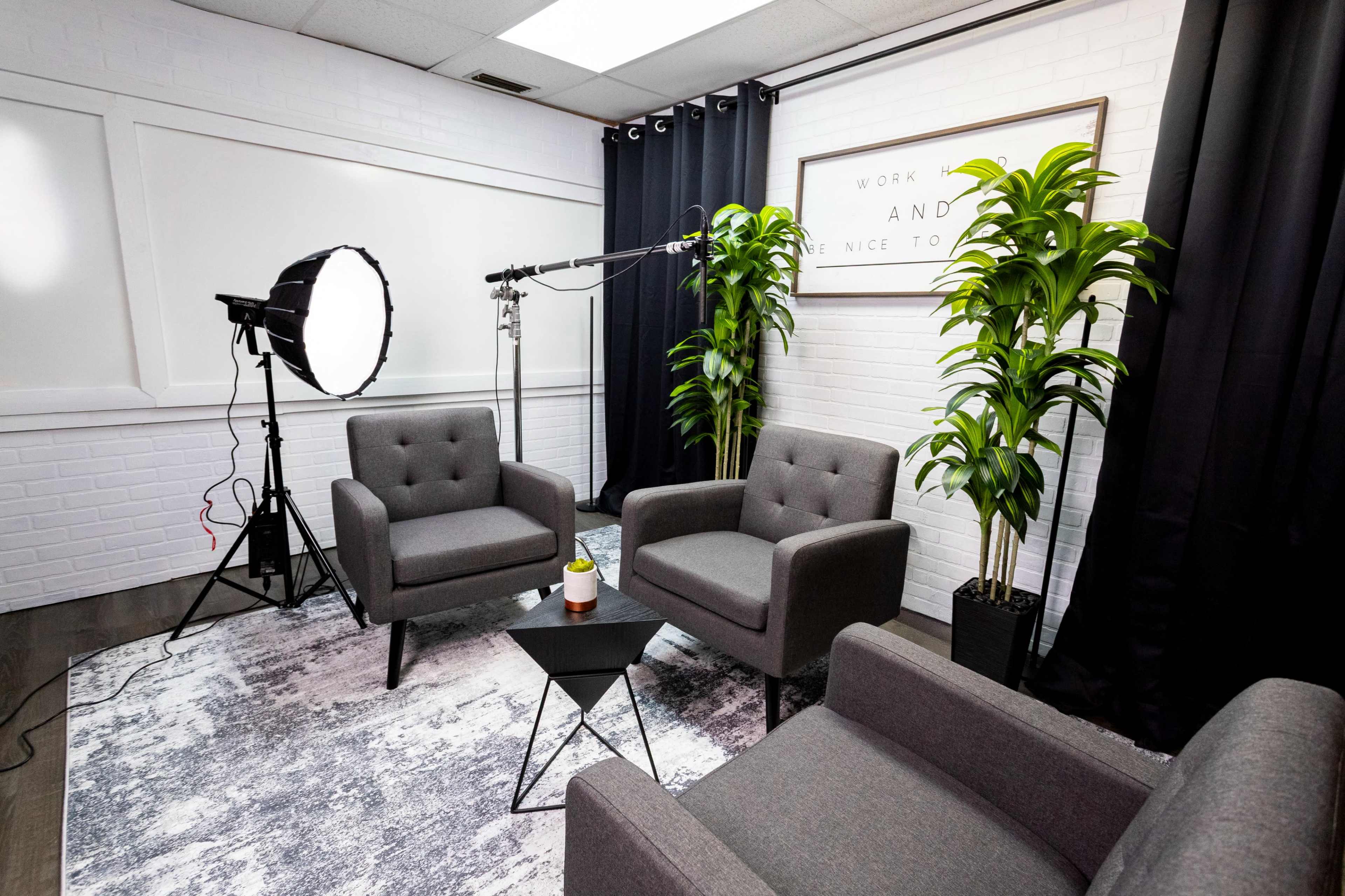 A bright room features three gray chairs arranged around a small black table, with a backdrop of plants and a lighting setup.