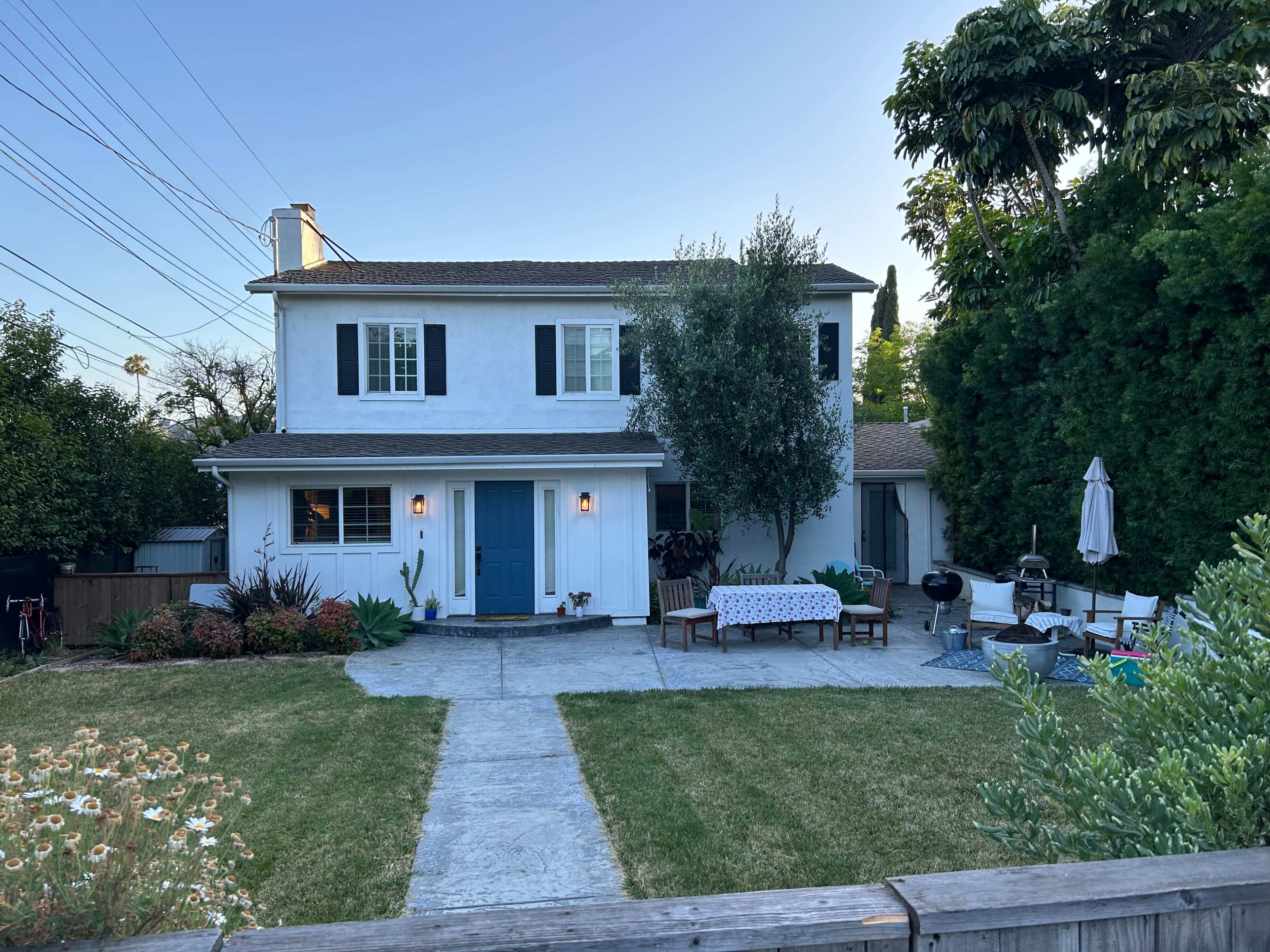 A two-story white house with blue doors and a landscaped yard features outdoor seating and various plants.