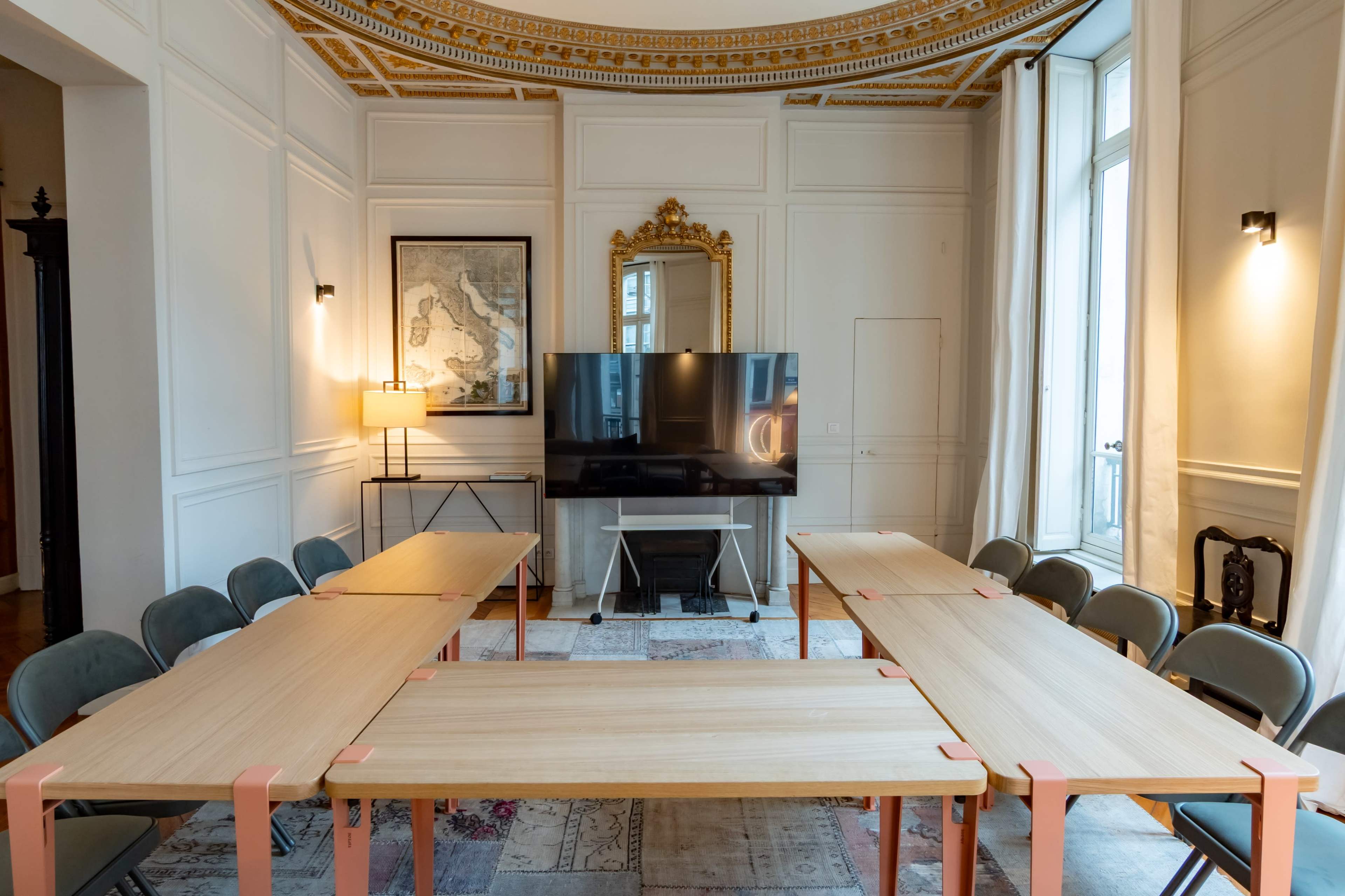 The image shows a modern meeting room with two rectangular tables arranged in a U-shape, a large television, and a lamp, all set against a backdrop of ornate ceilings and walls.