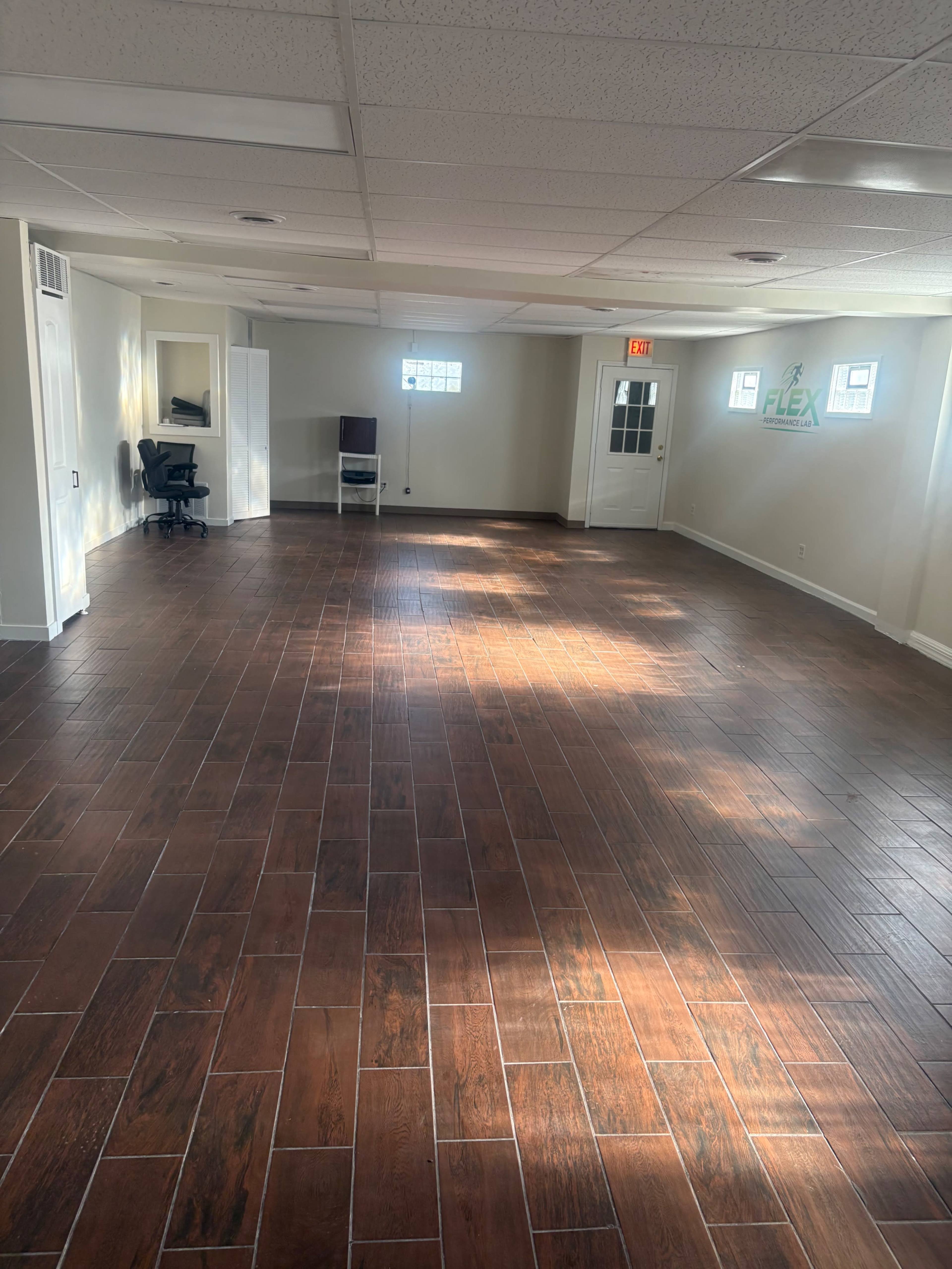 The image shows a spacious, empty room with hardwood flooring, a few chairs, and windows allowing natural light.