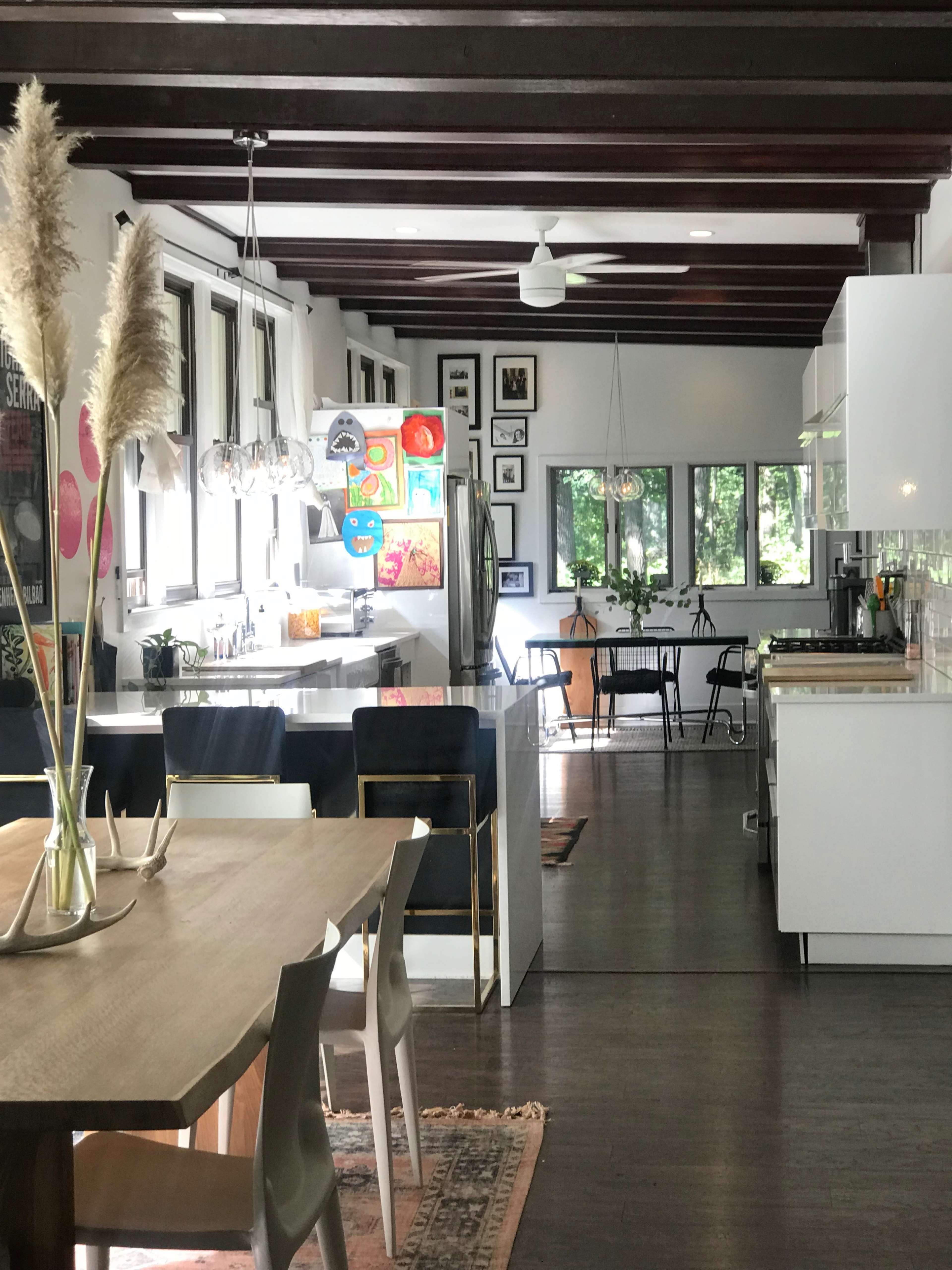 The image shows a modern, open-plan kitchen and dining area with wooden beams, a large dining table, and a variety of artwork on the walls.
