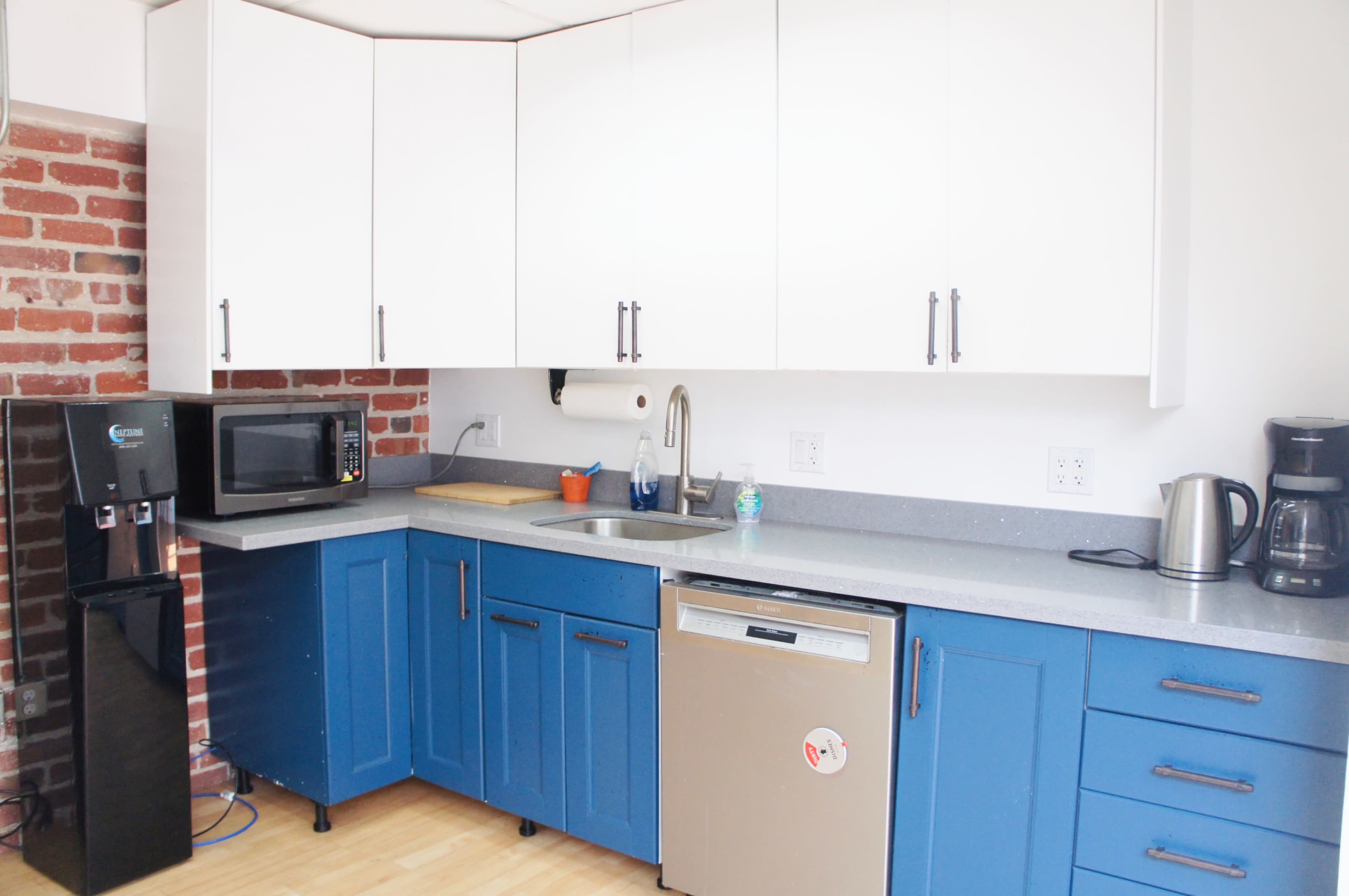 The image shows a modern kitchen with blue cabinets, a stainless steel dishwasher, a microwave, a coffee maker, and a brick wall.