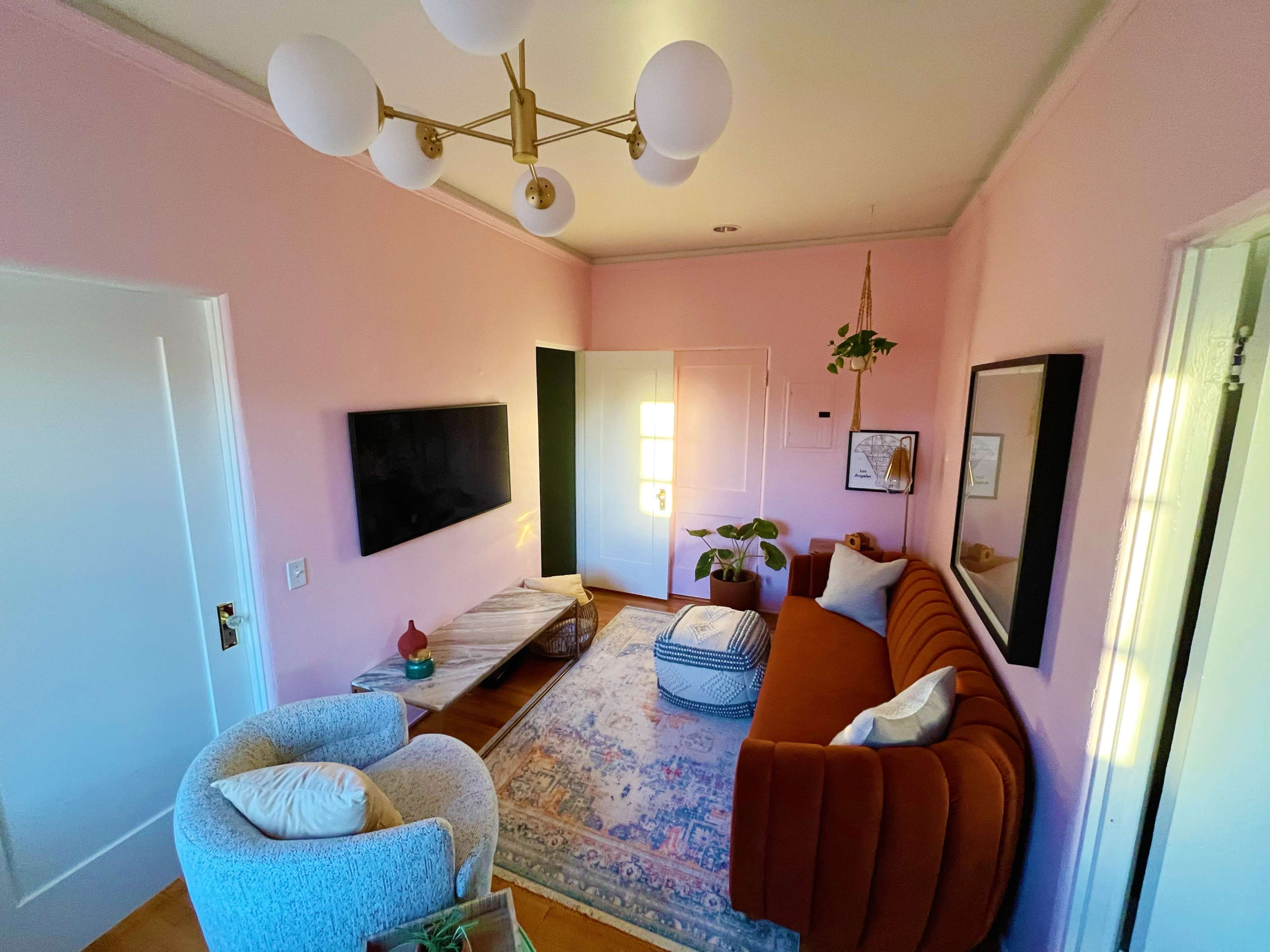 A living room with a pink wall, a brown sofa, a light fixture with multiple bulbs, a flat-screen TV mounted on the wall, and a decorative rug beneath a coffee table.
