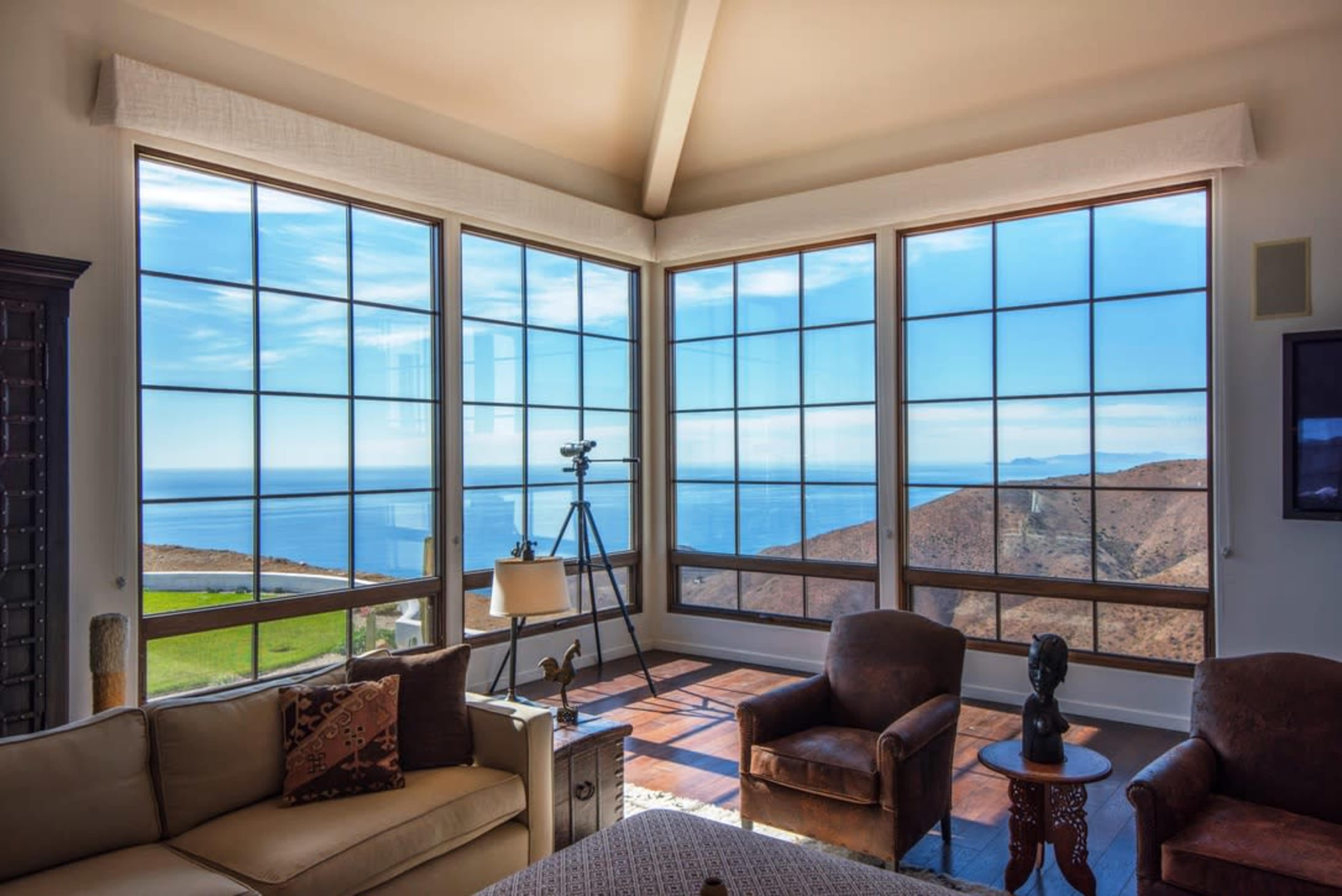 A spacious living room features large windows offering a view of the ocean and hills, with a telescope positioned near the glass.