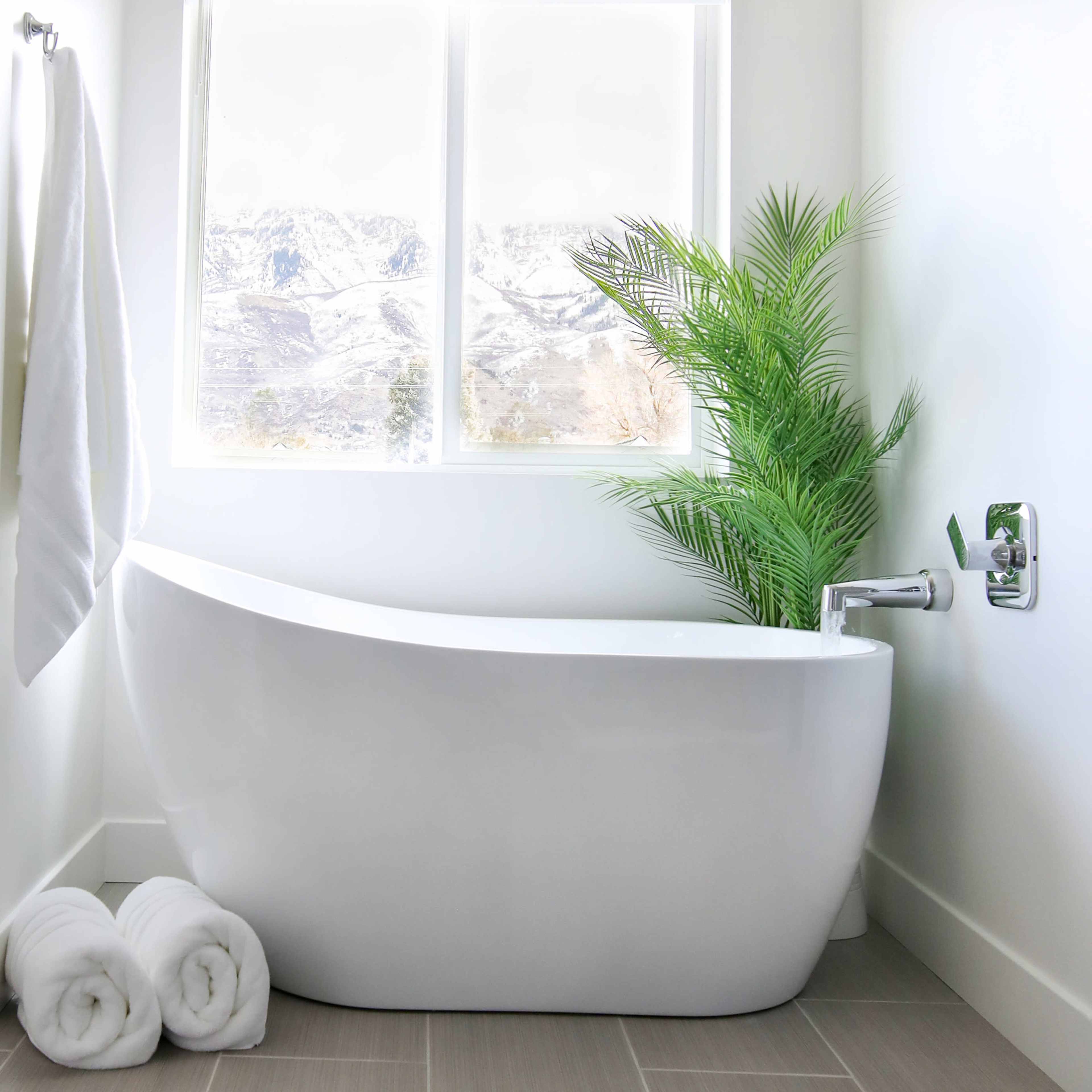 A modern bathroom features a freestanding white bathtub beside a large window with a view of snow-covered mountains, complemented by a potted plant and neatly rolled towels.