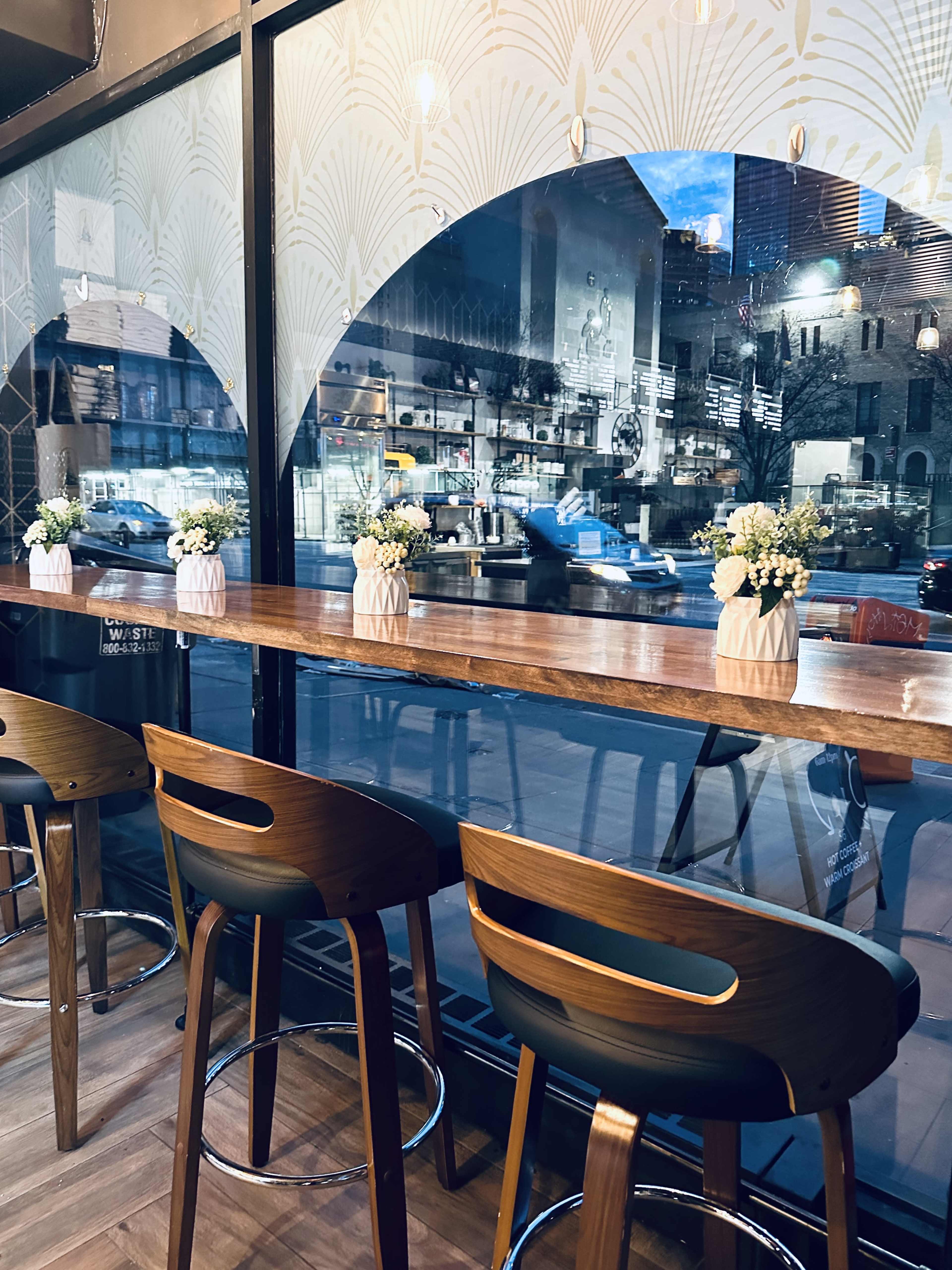 The image shows a modern café interior with wooden bar stools lined up at a counter, facing large windows that overlook a street.