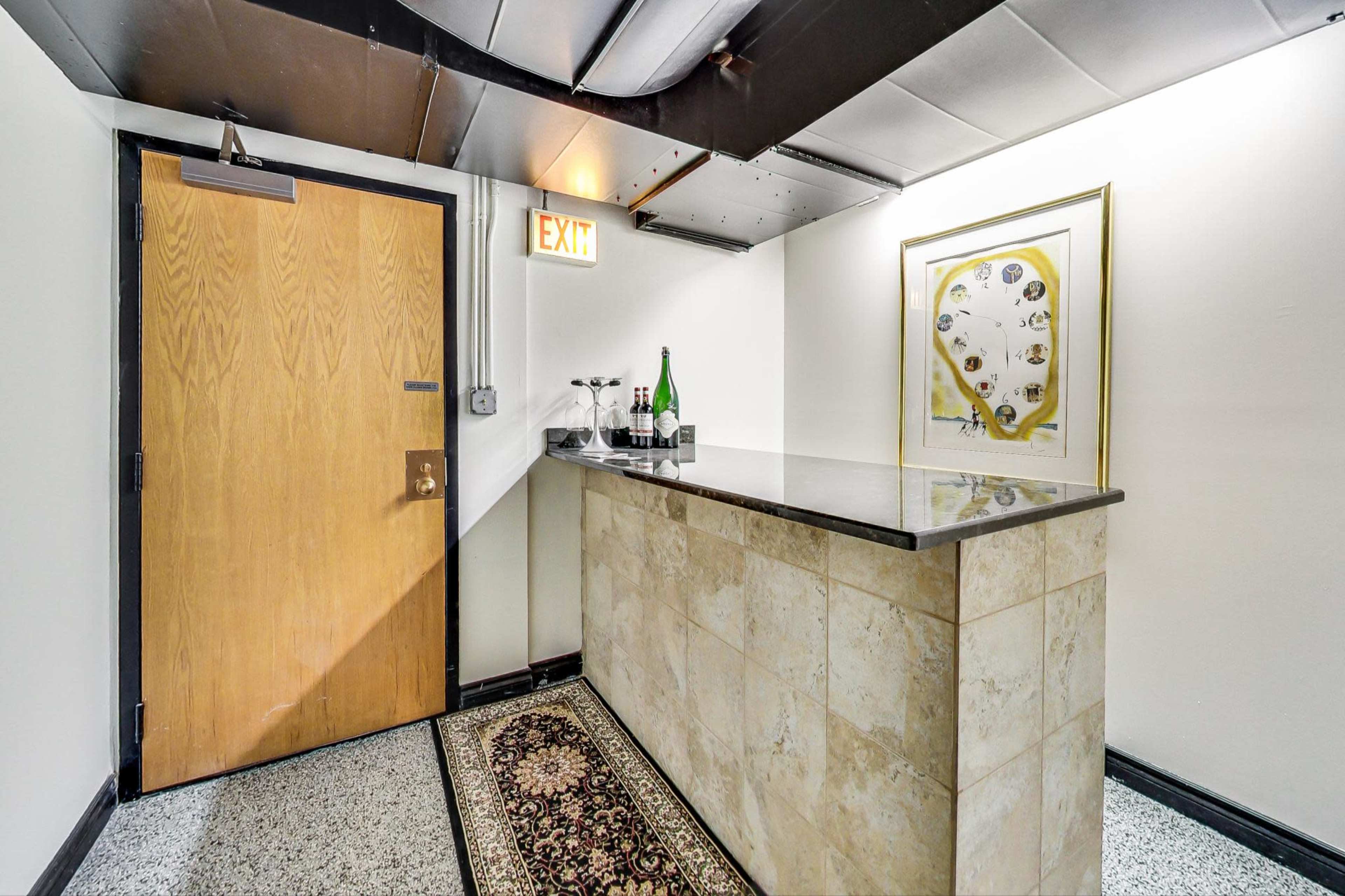 A small hallway features a wooden door, an exit sign, and a bar with a stone countertop displaying bottles and glasses.
