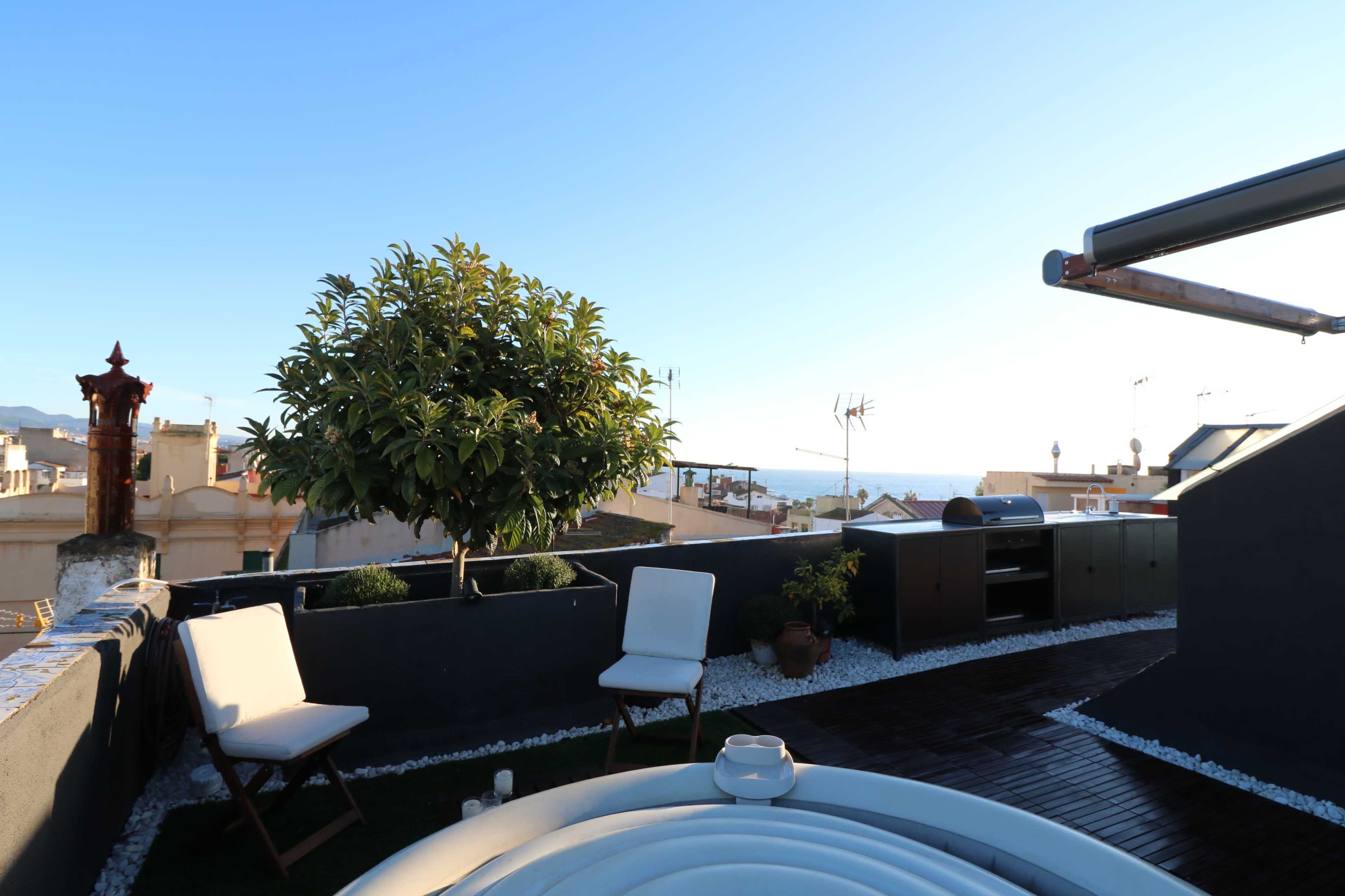 The image shows a rooftop terrace with two chairs, a small tree, and a view of the ocean in the background.