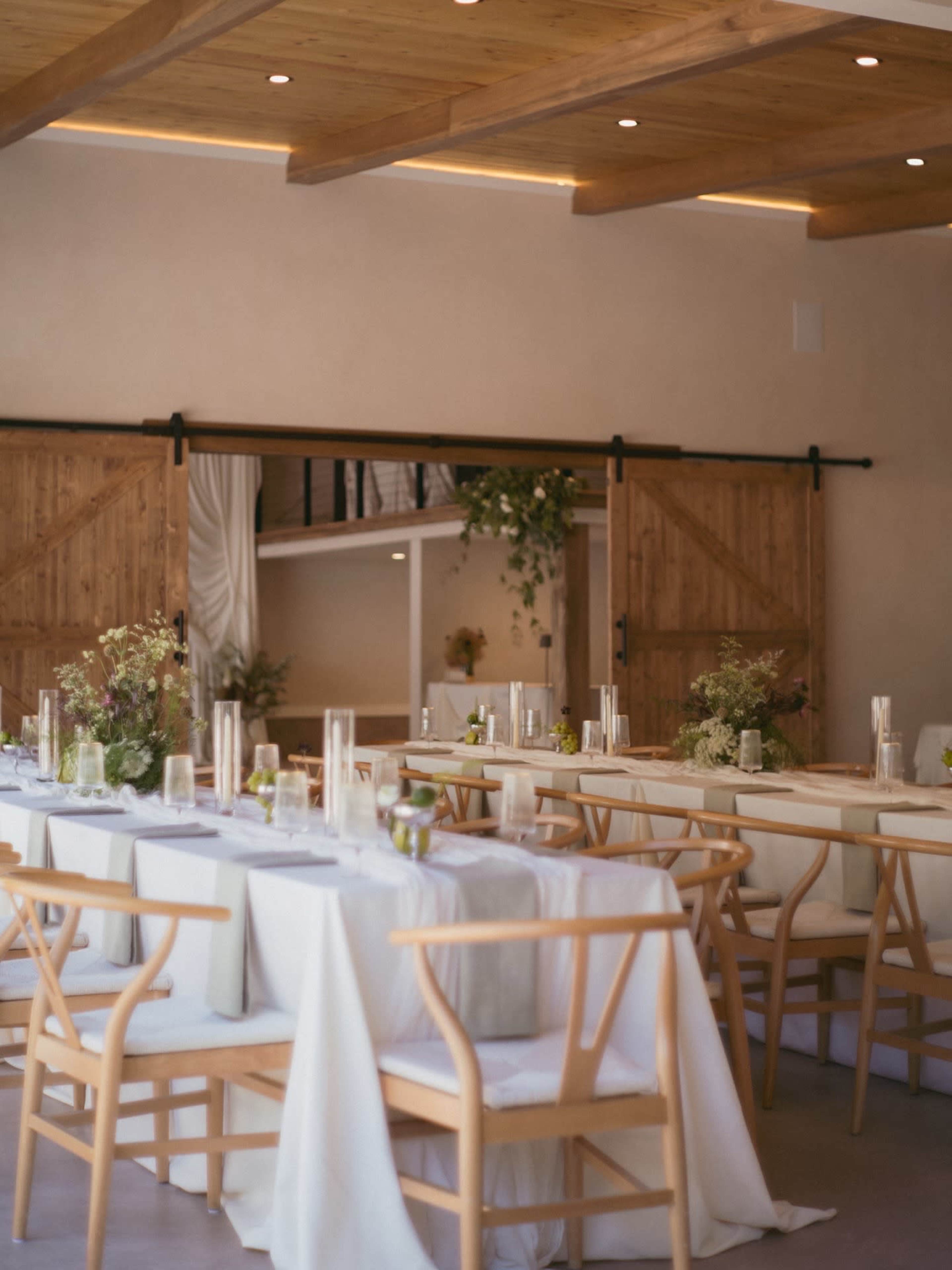 A long dining table is set with simple centerpieces and surrounded by wooden chairs in a softly lit room with wooden beams and sliding barn doors.