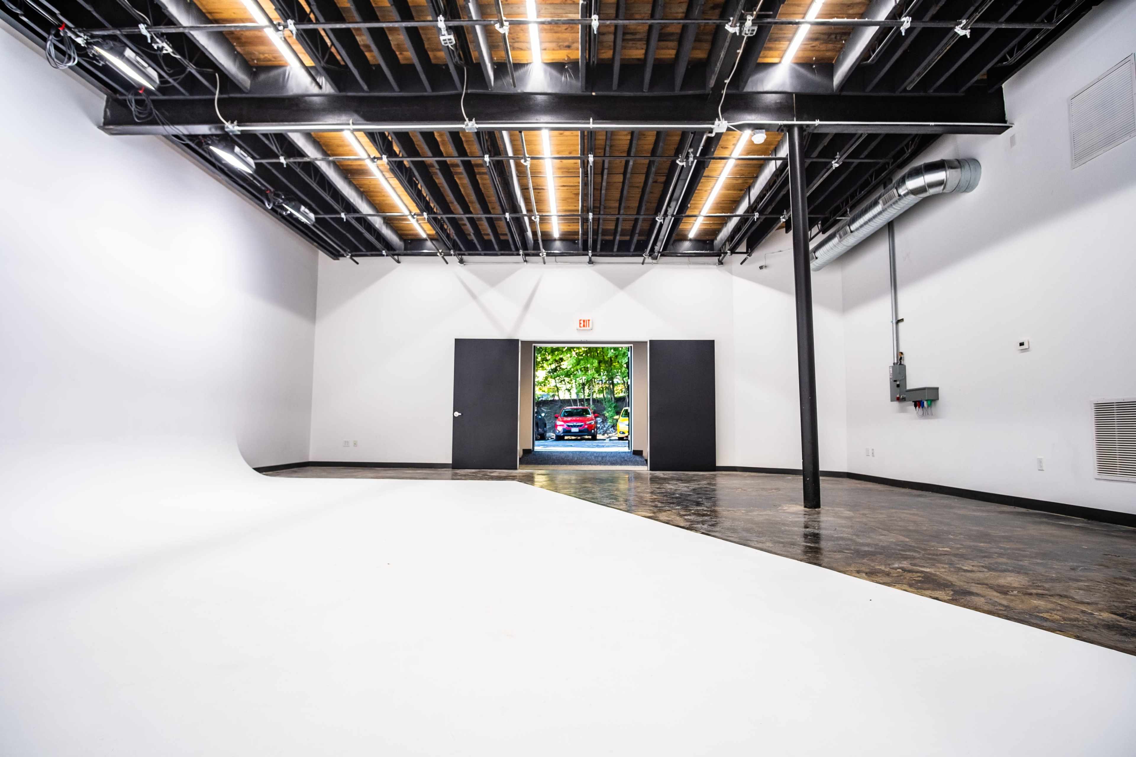 An empty studio space with a curved white surface and a view of a car parked outside through double doors.