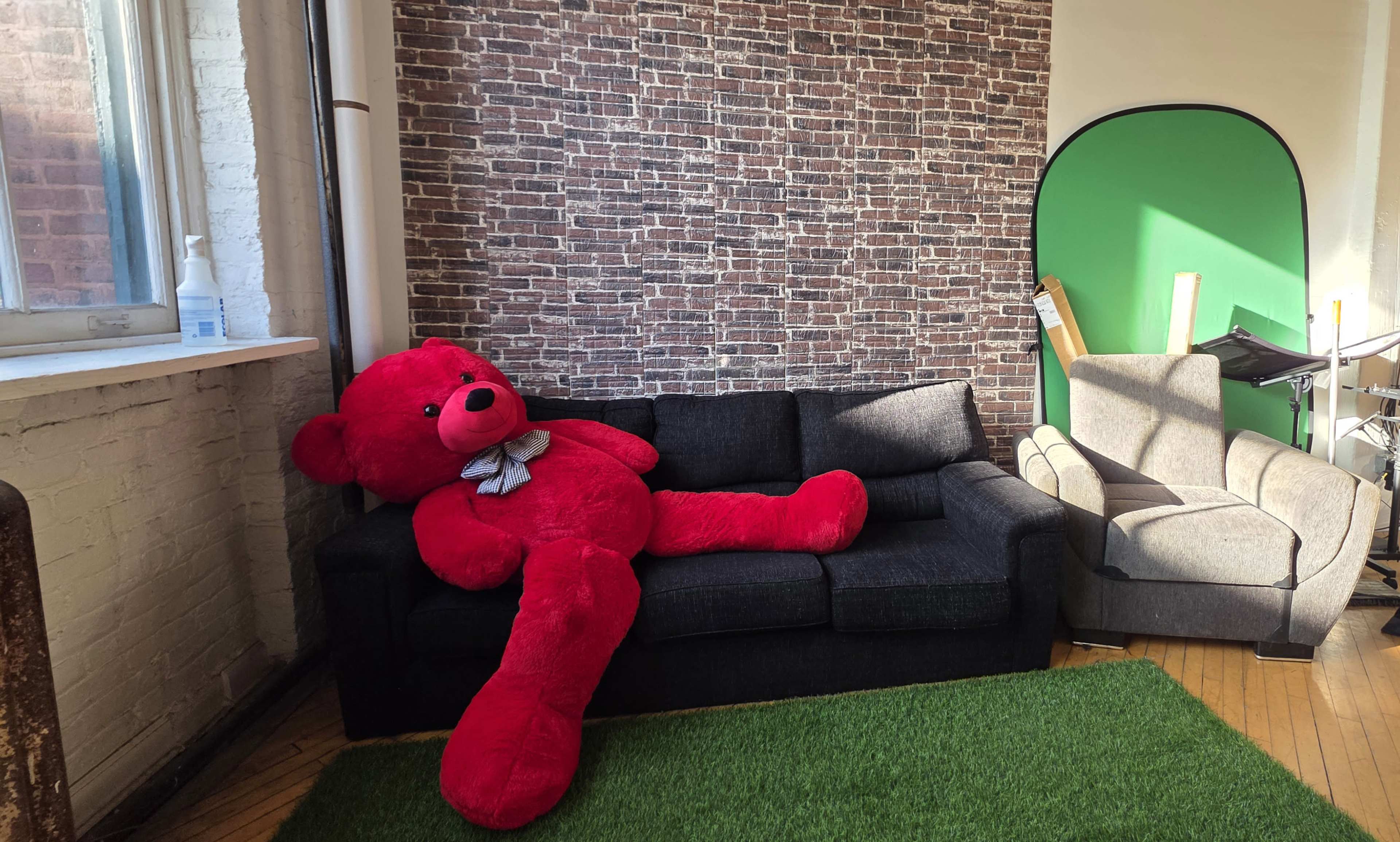 A large red teddy bear is leaning on a dark sofa in a room with a brick-patterned wall and green backdrop.