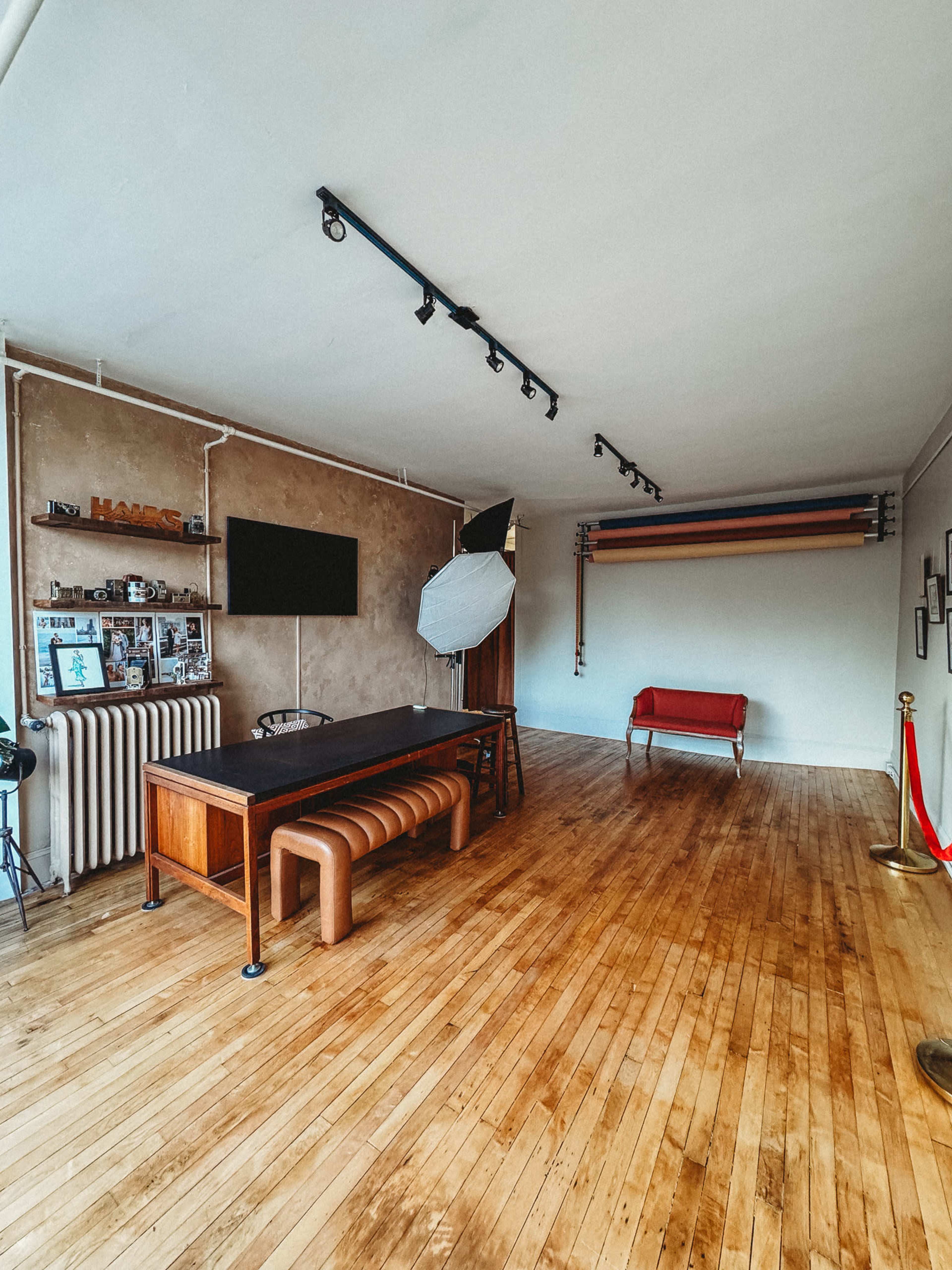 The image shows a spacious room with wooden flooring, featuring a long table, a red bench, a wall-mounted television, and lighting fixtures above.