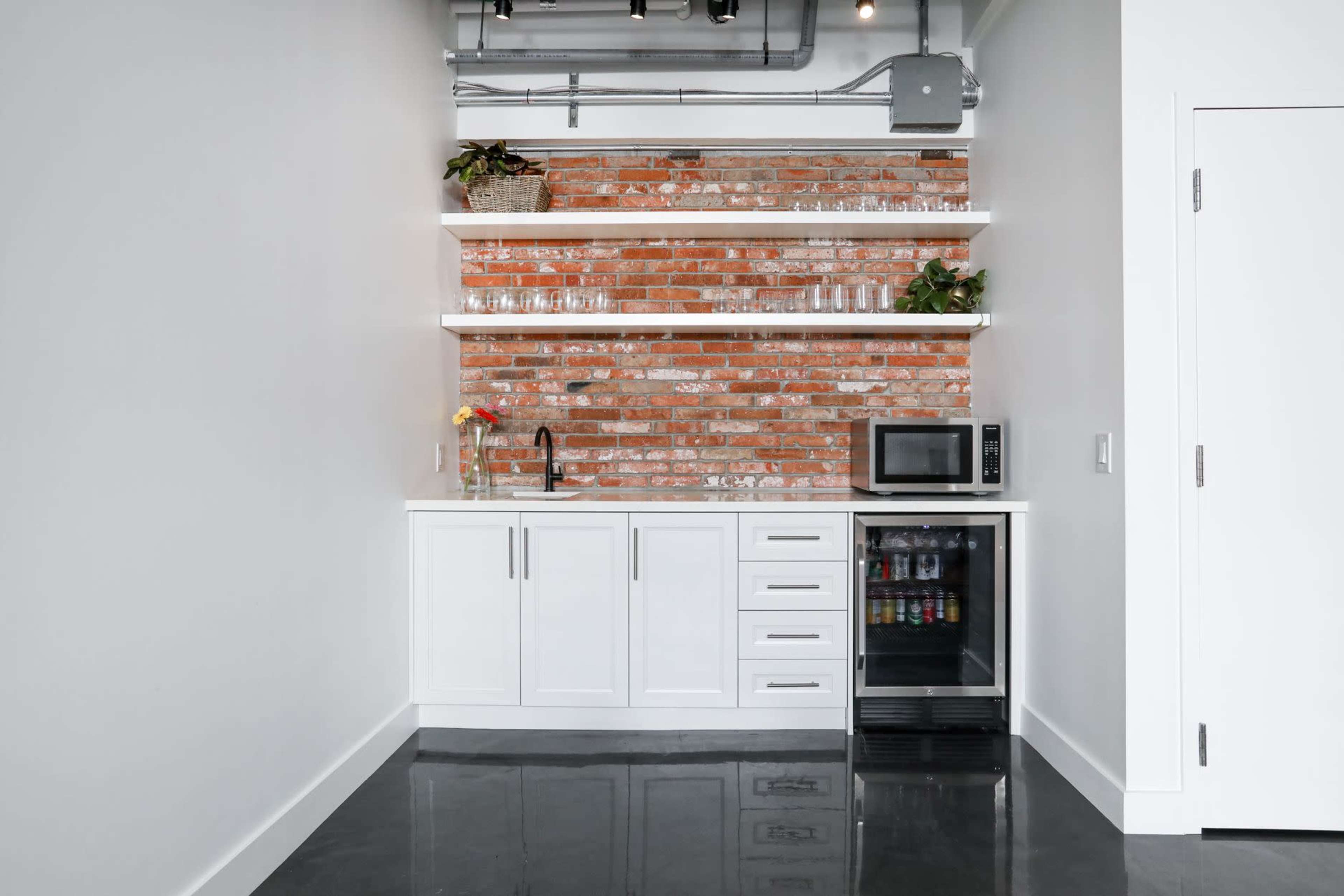 A modern kitchenette featuring a brick wall, white cabinetry, open shelves, a sink, a microwave, and a mini refrigerator.
